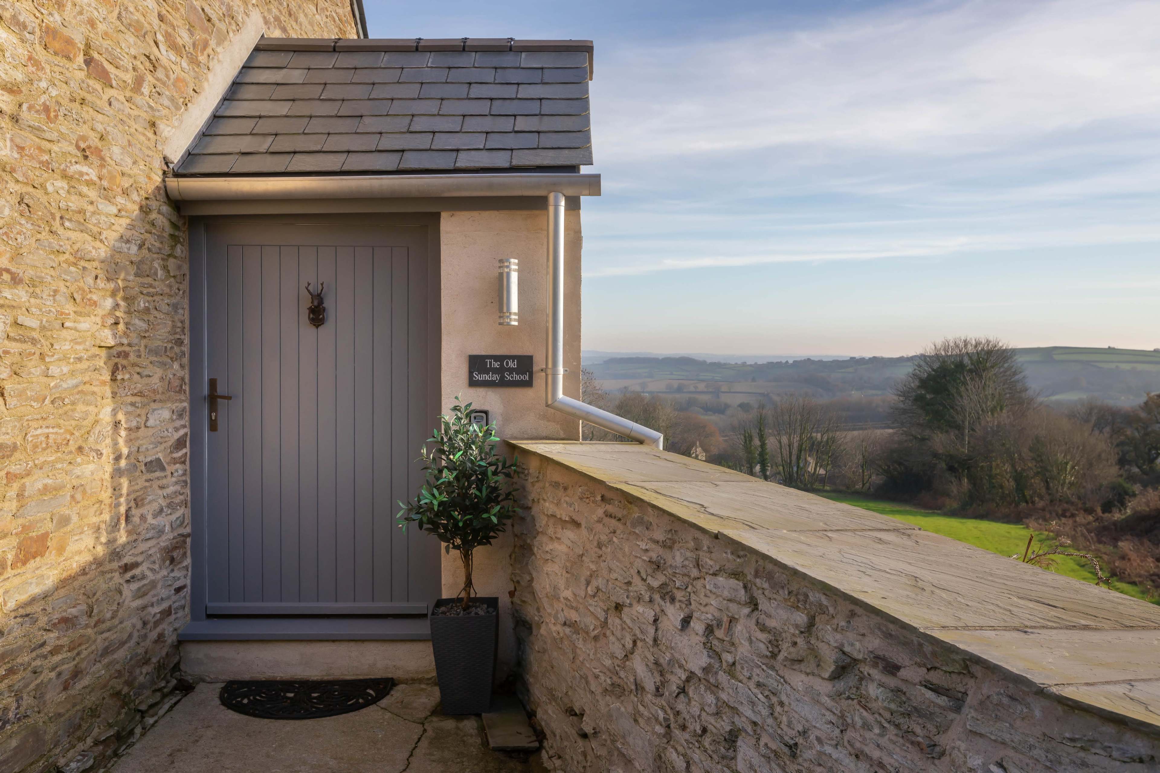 The entrance of a building with a gray door and a sign that reads "The Old Smiddy School," set against a backdrop of rolling hills and clear blue skies.
