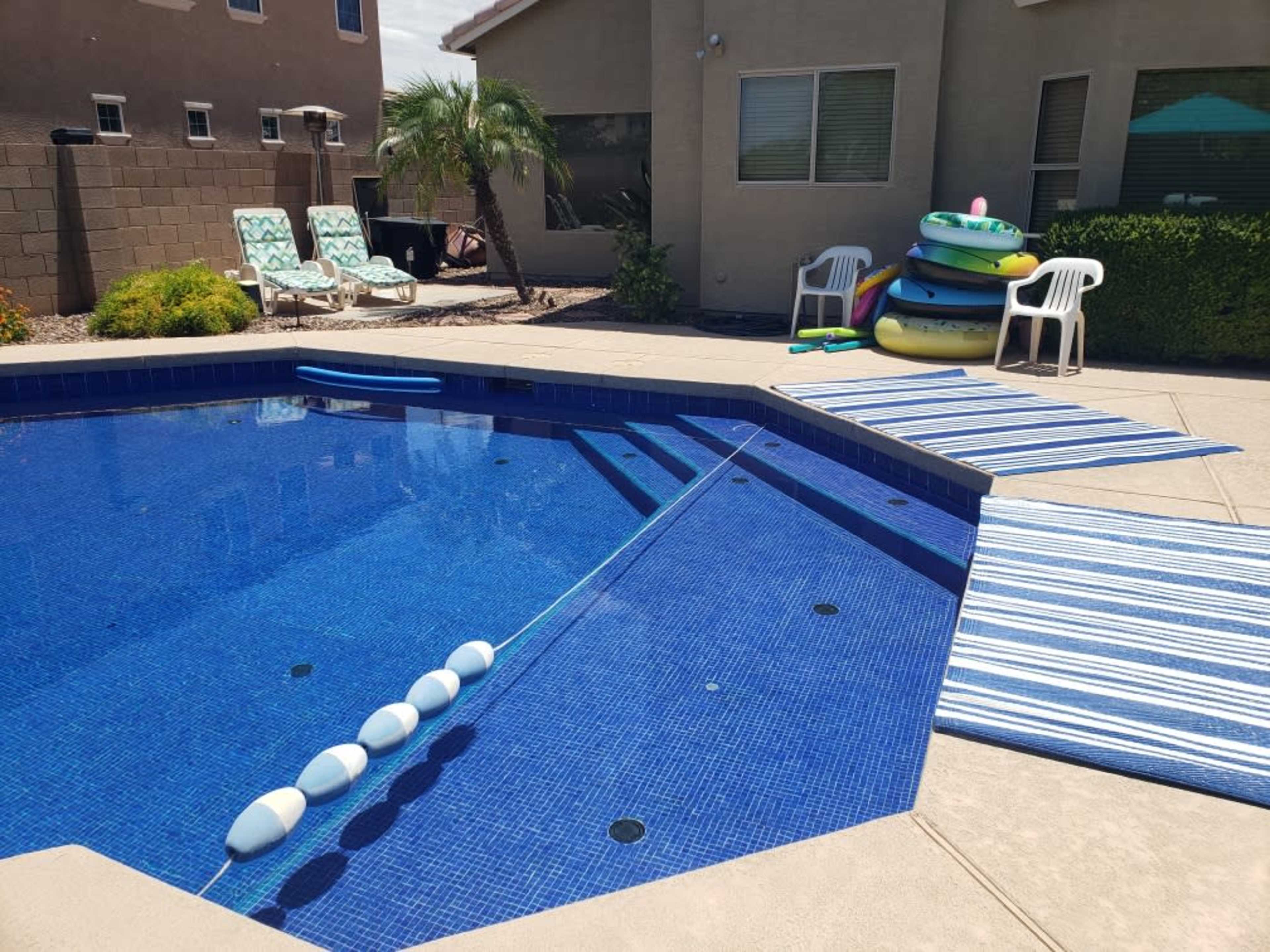 A blue-tiled swimming pool is bordered by lounge chairs, a stack of colorful inflatable rafts, and a striped mat leading to a patio.