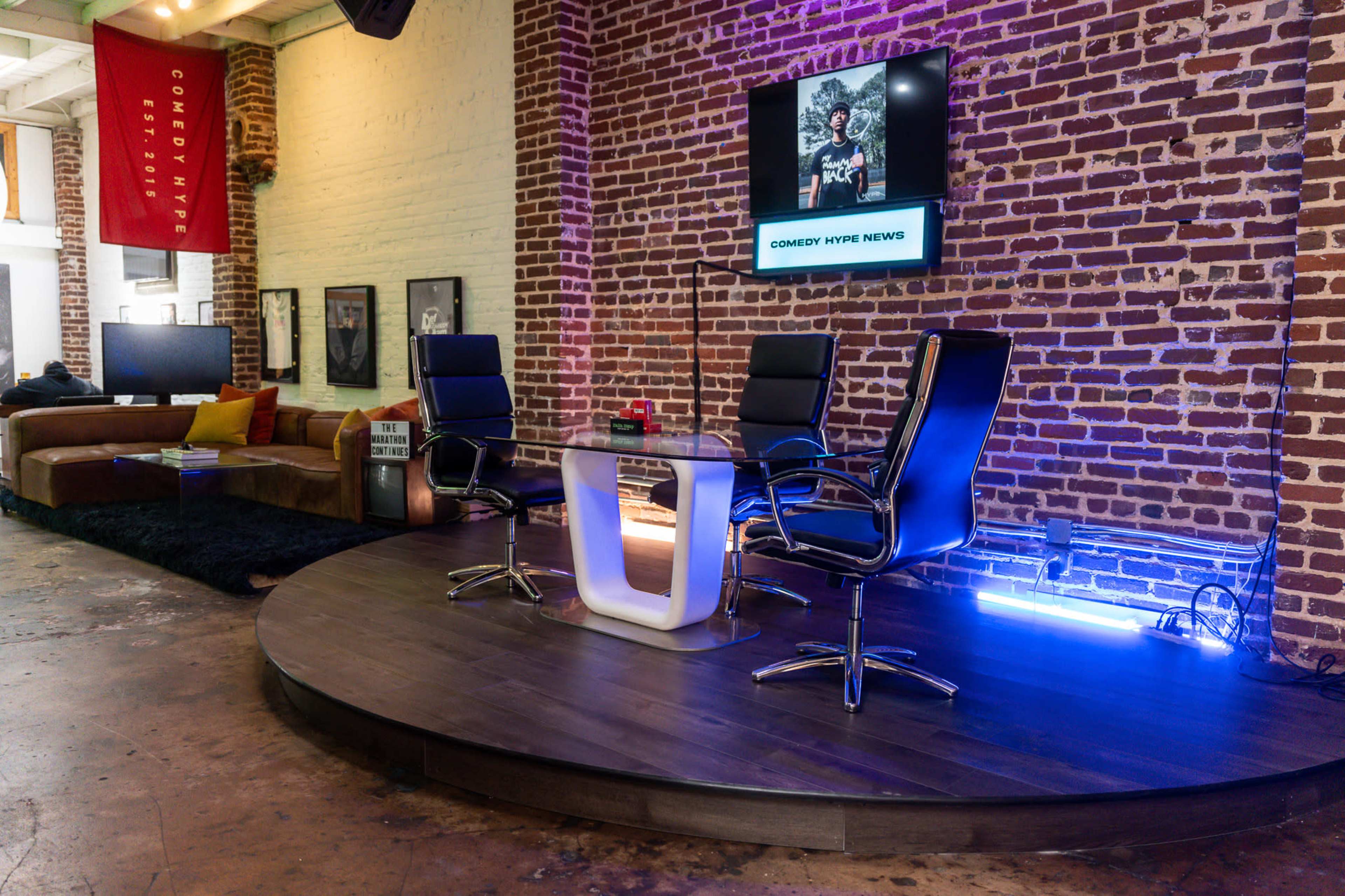 The image shows a modern studio setup with a circular wooden platform, three black chairs, and a television displaying "Comedy Hype News" against a brick wall backdrop.