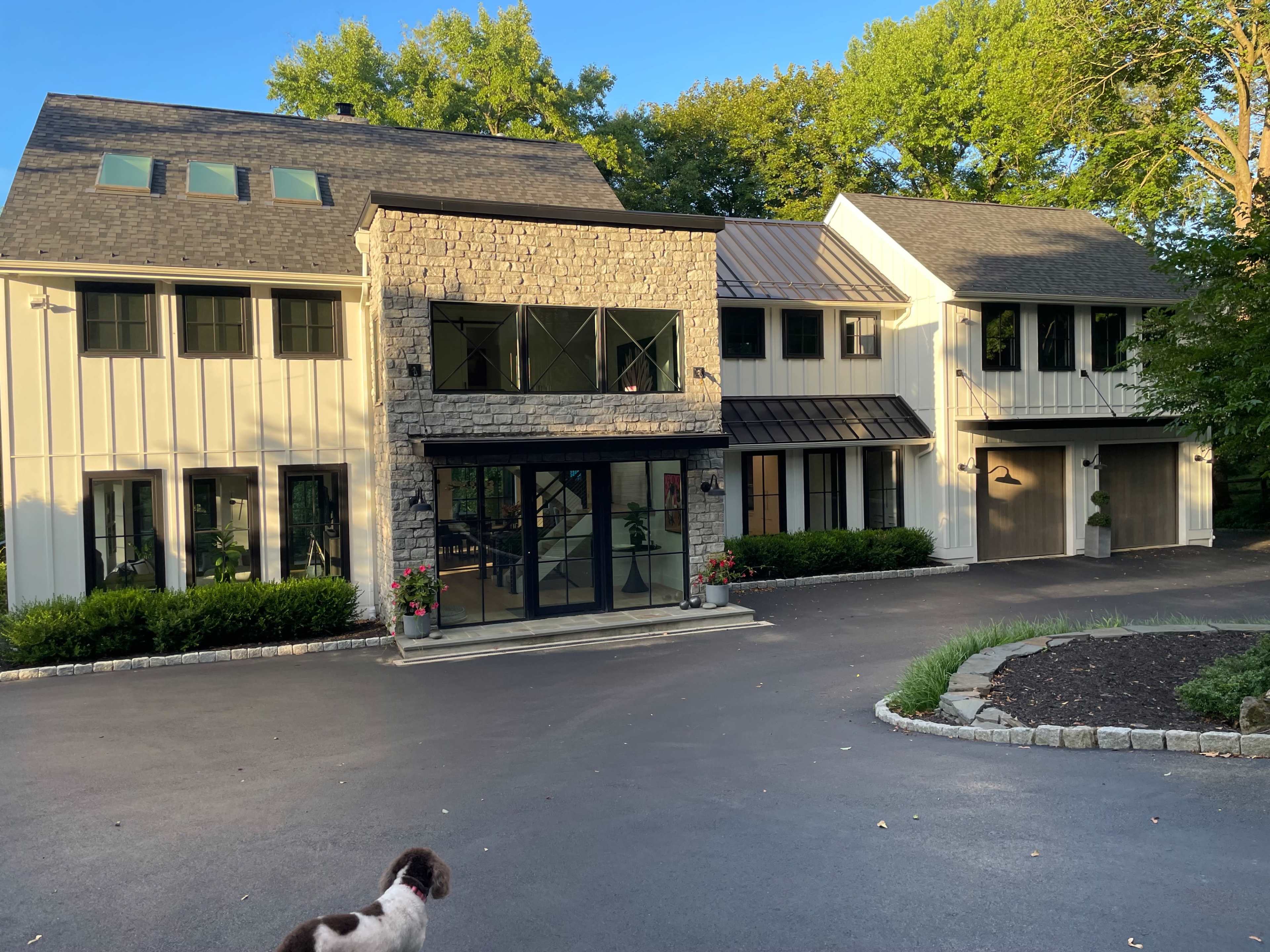 A modern two-story house features a central stone facade, large glass windows, and a two-car garage, surrounded by a landscaped driveway.
