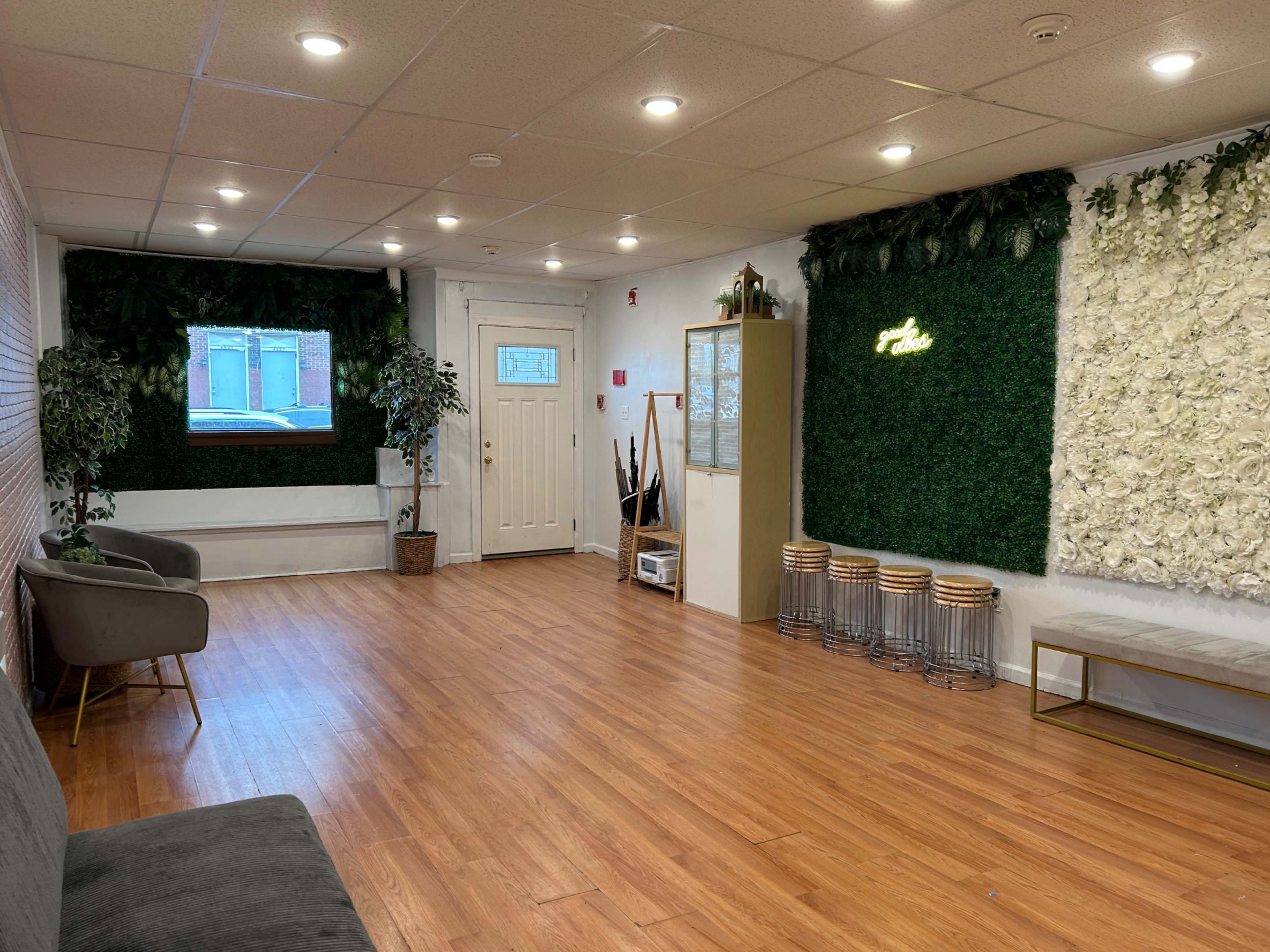 The image shows a spacious, well-lit interior room featuring a wooden floor, green wall decorations, and modern furniture arranged for a welcoming atmosphere.