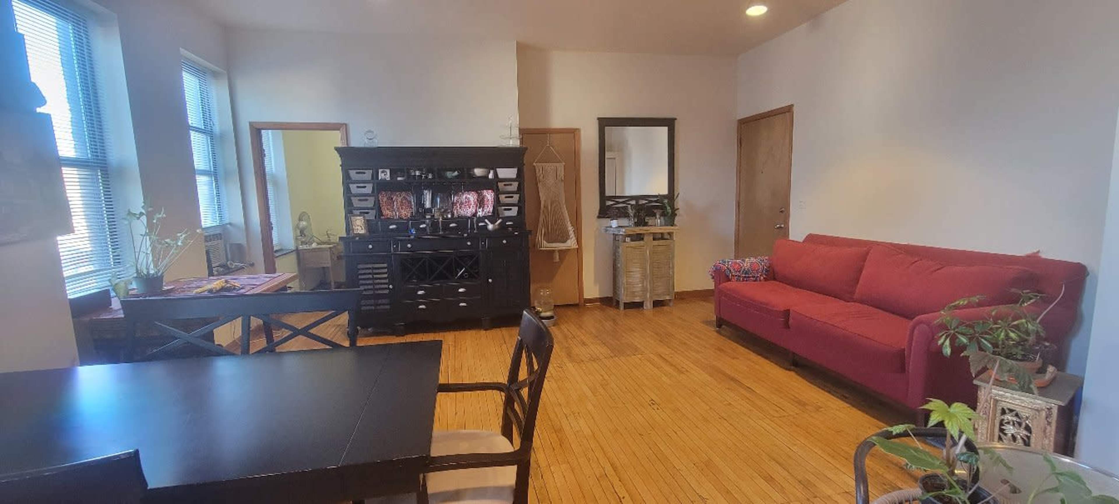 A living space featuring a red couch, a dining table, a black cabinet, and wooden flooring with several decorative items.