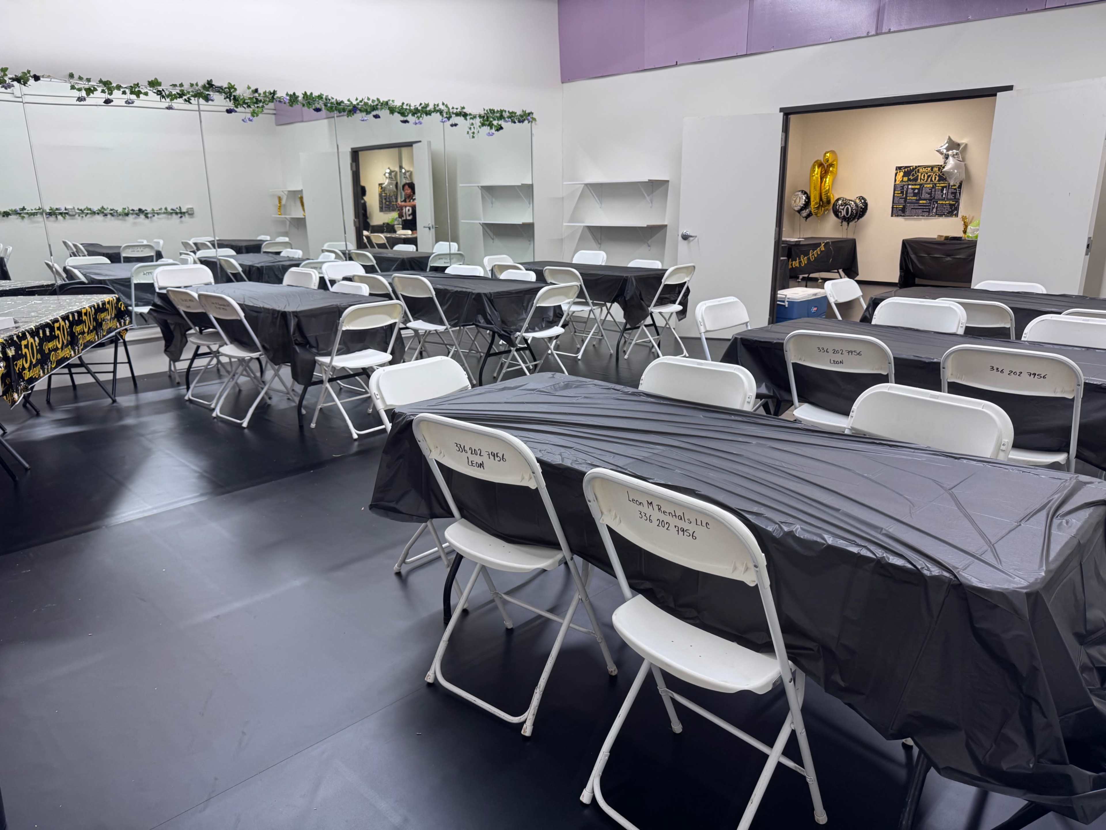 The image shows a room set up with numerous black tables covered with tablecloths and white folding chairs arranged for an event.