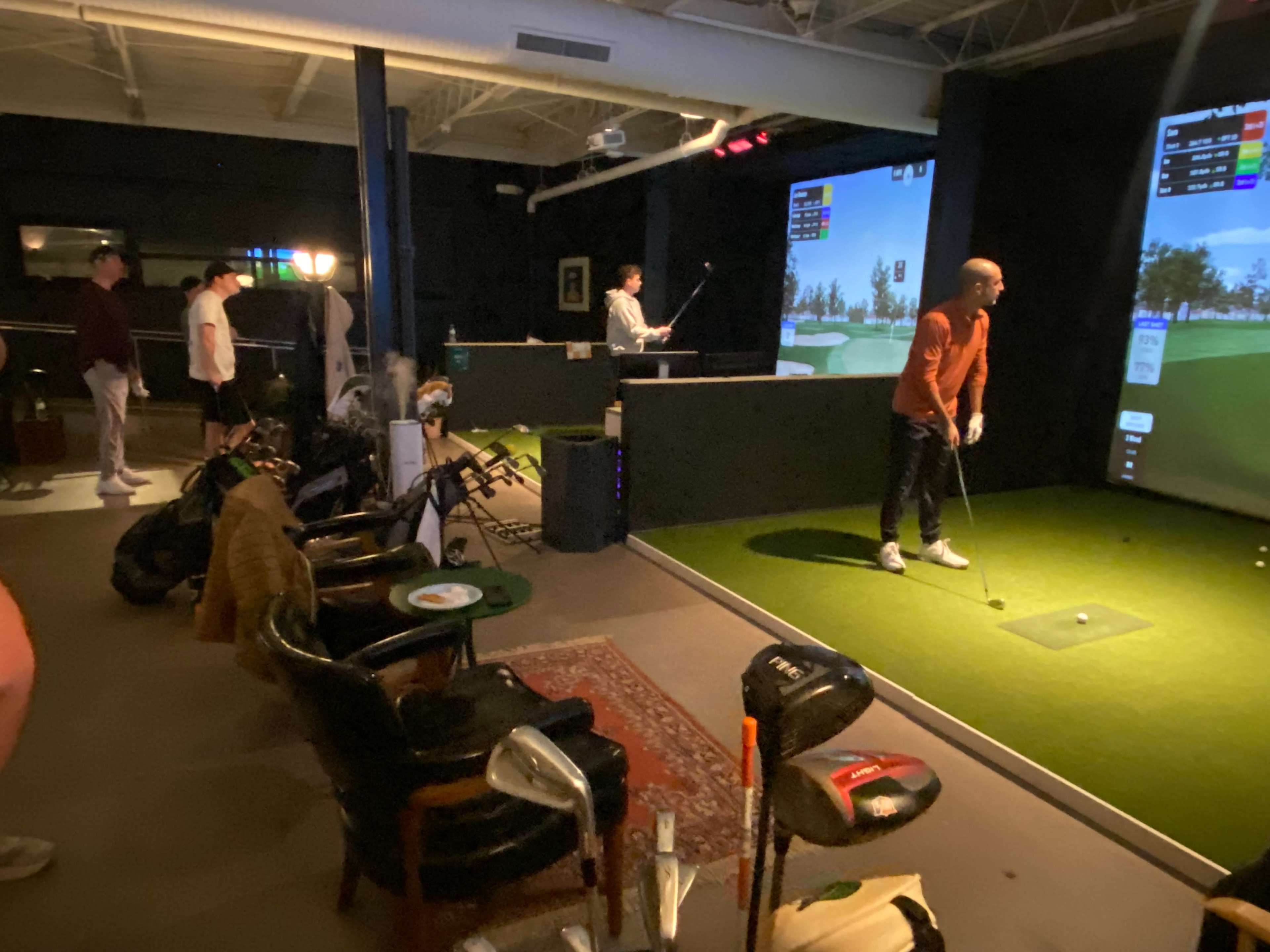 The image shows a group of people in an indoor golf simulator facility, with one man preparing to hit a golf ball while others observe.