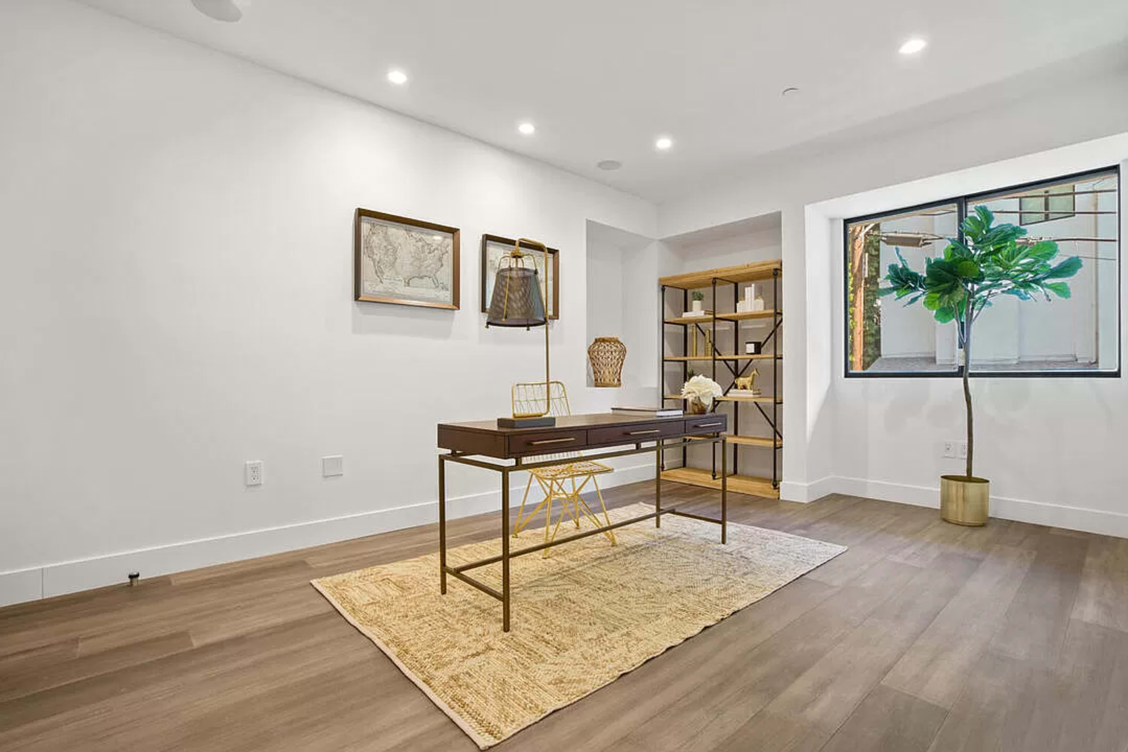 A modern home office featuring a wooden desk with a lamp, a bookshelf, and a potted plant, set against a bright white wall.