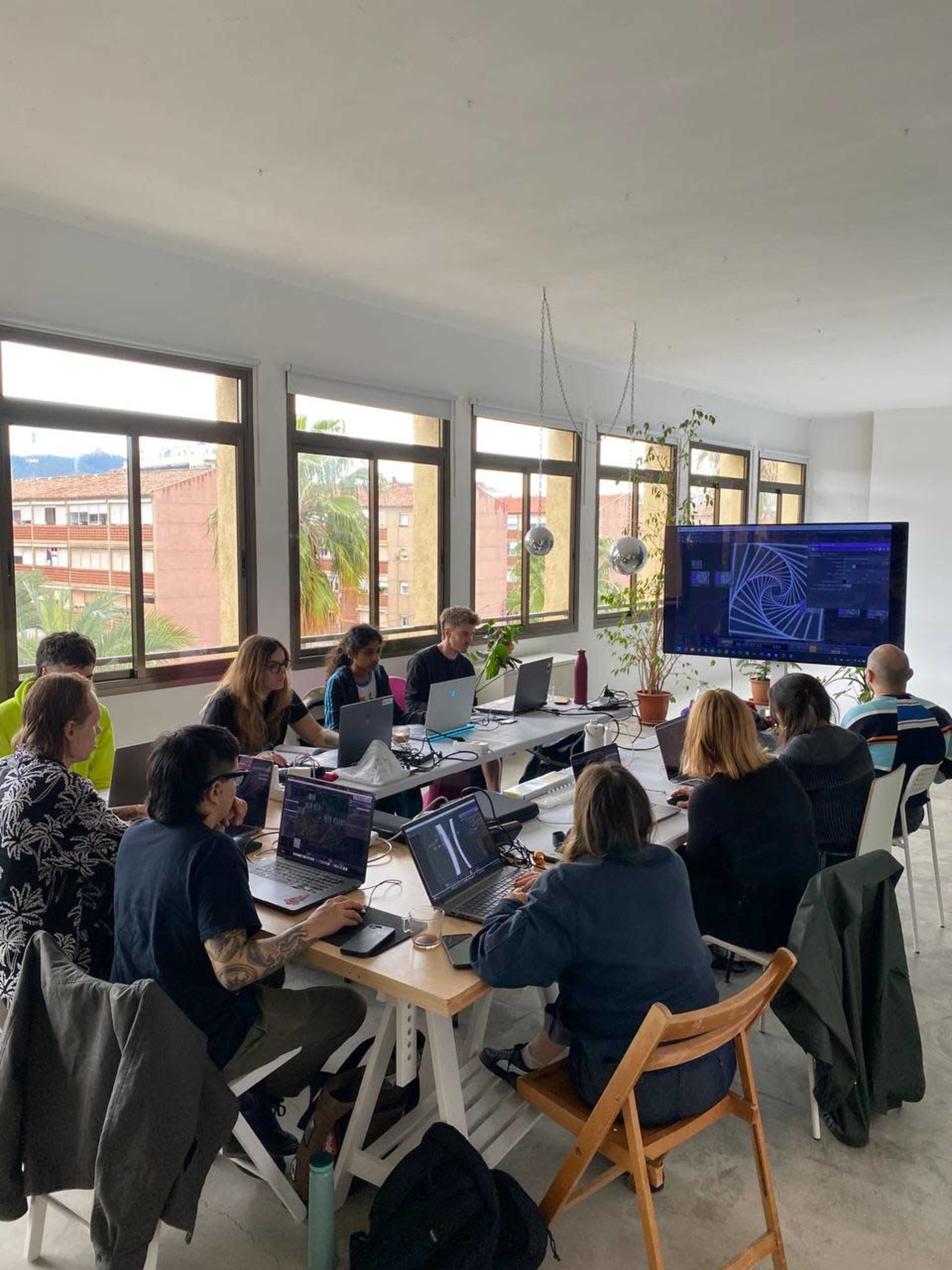 A group of individuals sits at a long table in a bright room, working on laptops while facing a large screen displaying graphics.