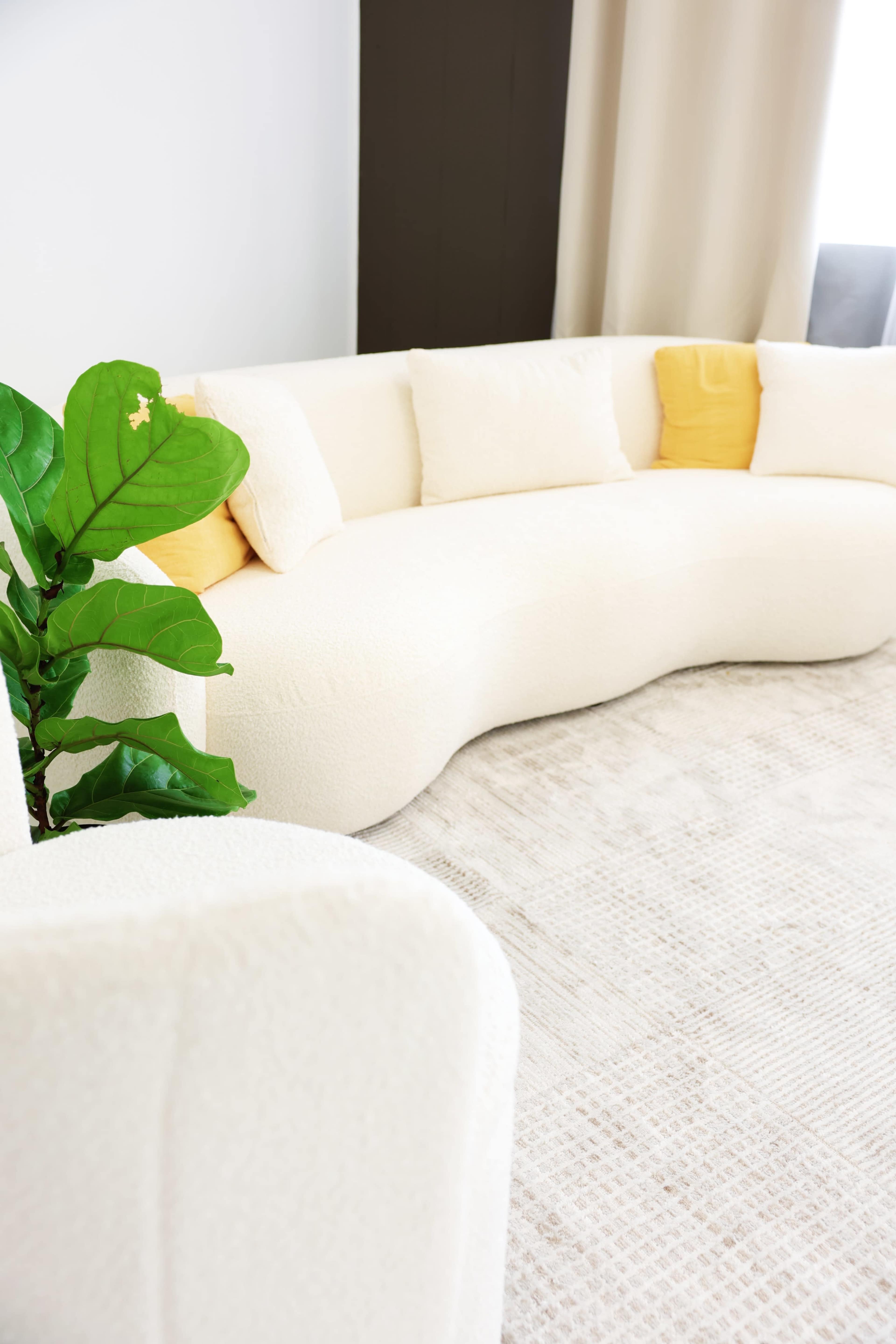 A curved white sofa with yellow accent pillows is positioned next to a potted plant on a light-colored rug.
