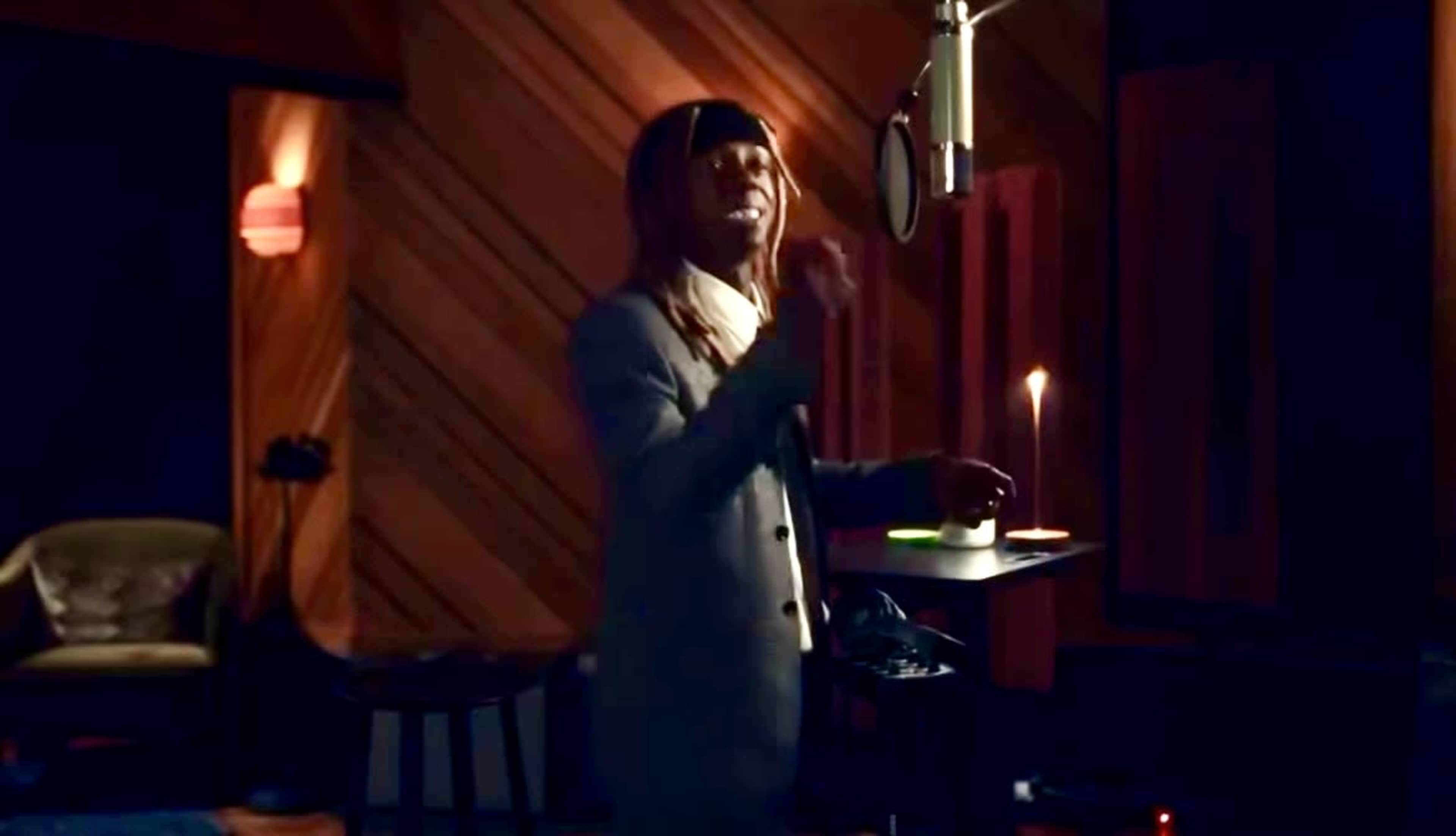 A person stands in a recording studio, speaking into a microphone with a candle placed on a small table beside them.