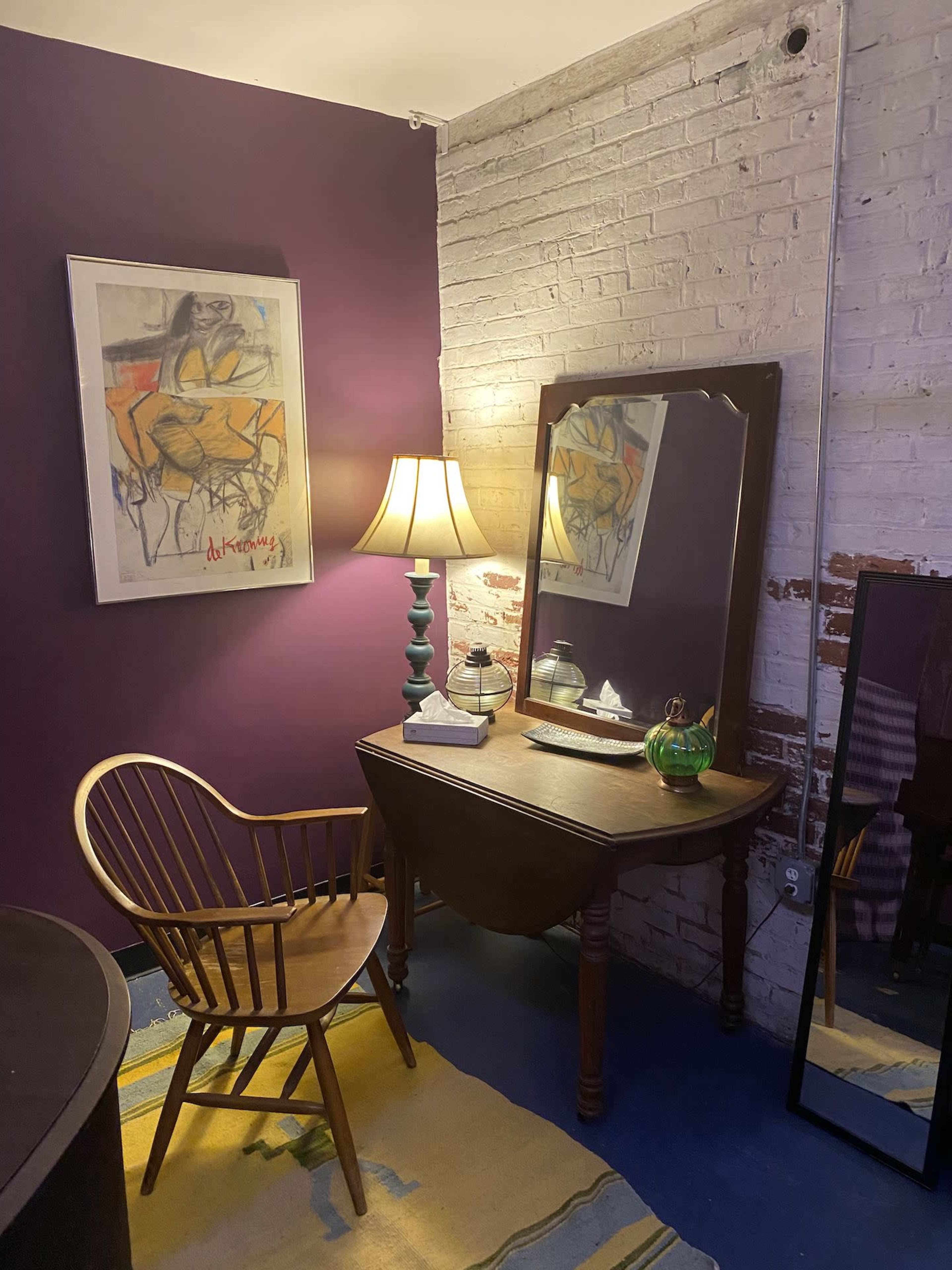 A corner of a room featuring a wooden chair, a dark wooden vanity with a mirror, a lamp, and a colorful rug against a purple wall and exposed brick.