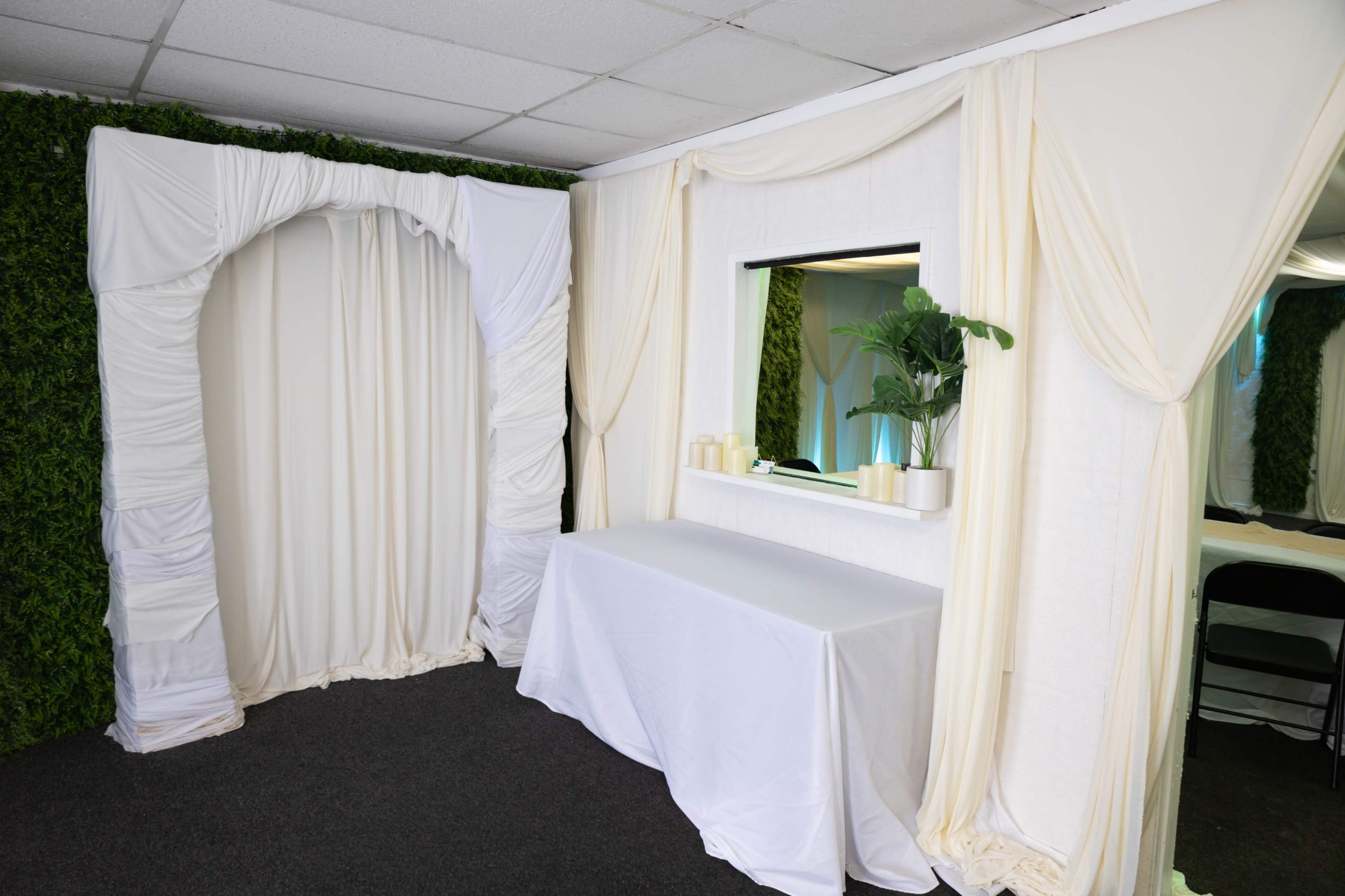The image shows a small room decorated with white drapes and a mirror, featuring a table and a potted plant against a backdrop of green foliage.