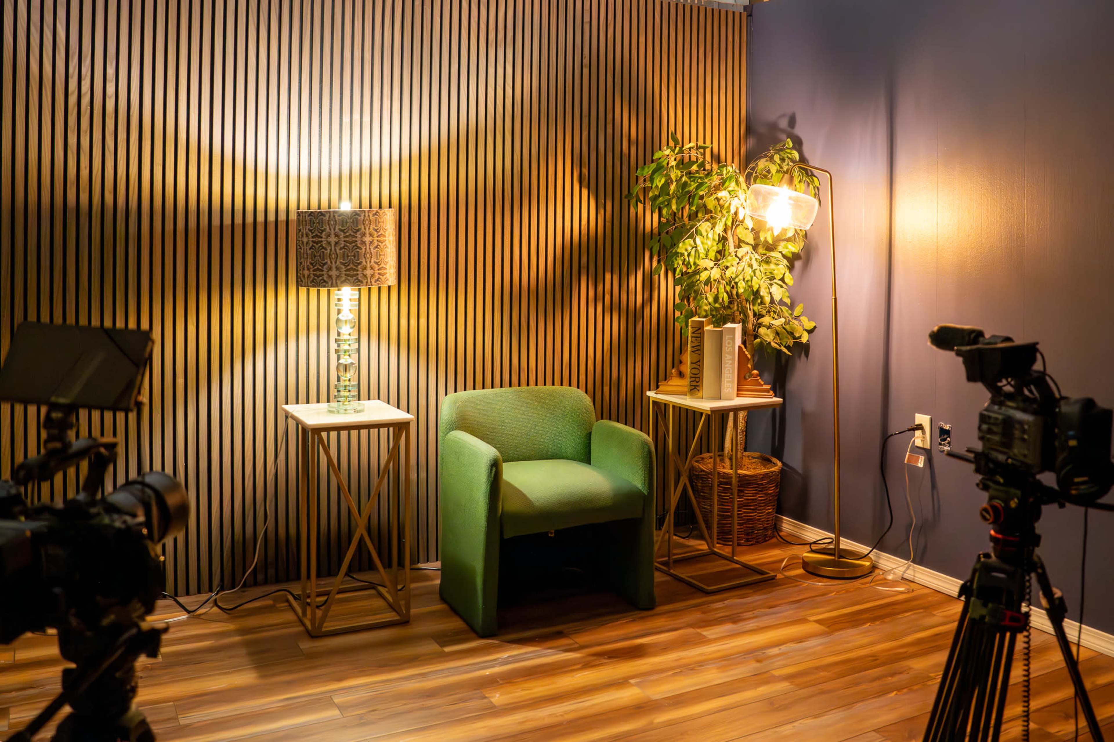 The image depicts a small, well-lit studio setup featuring a green chair flanked by two side tables, a lamp, a plant, and multiple cameras positioned in the foreground.