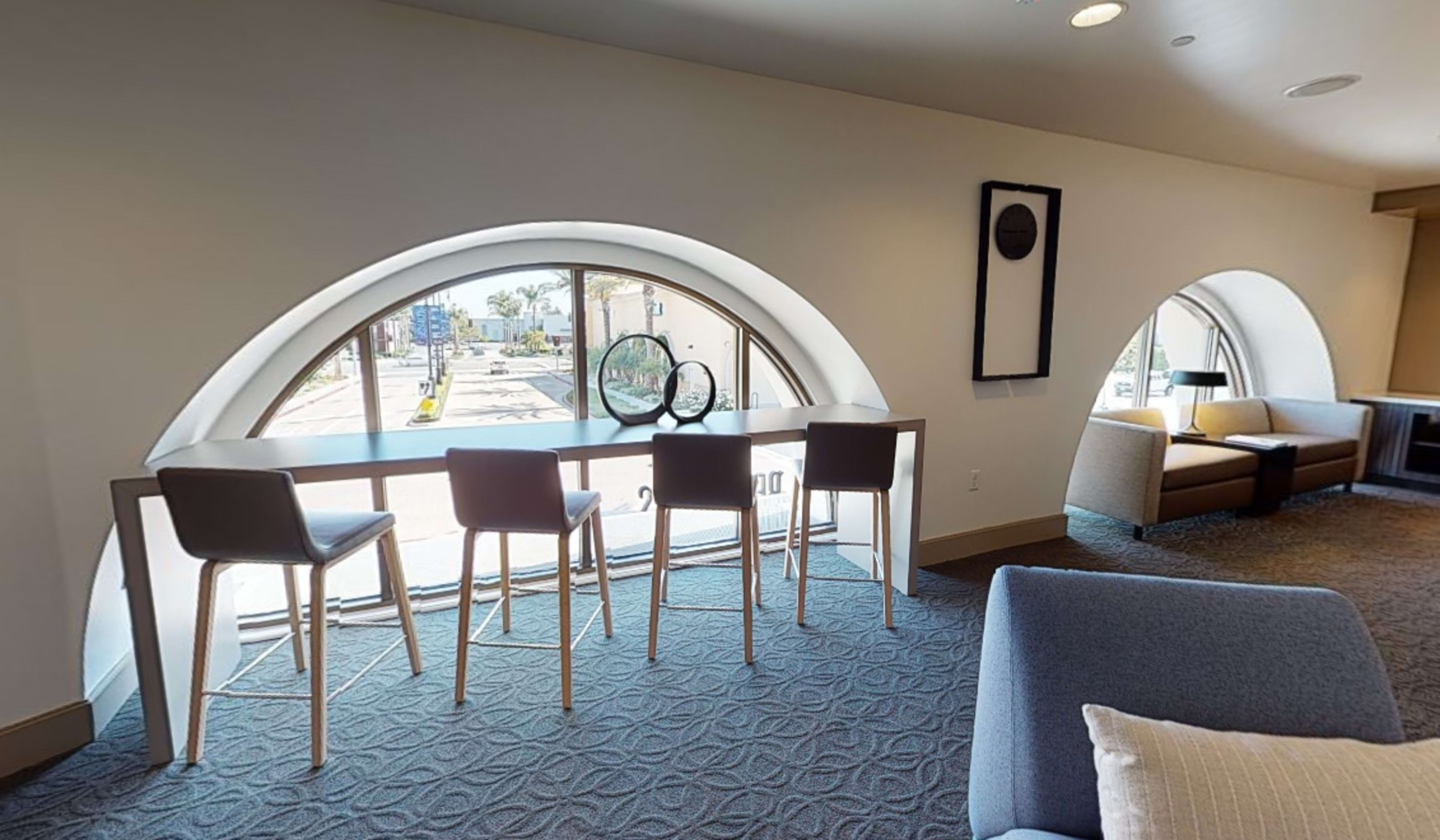 A modern interior space with a long table and four high chairs positioned in front of large arched windows, overlooking an outdoor area.