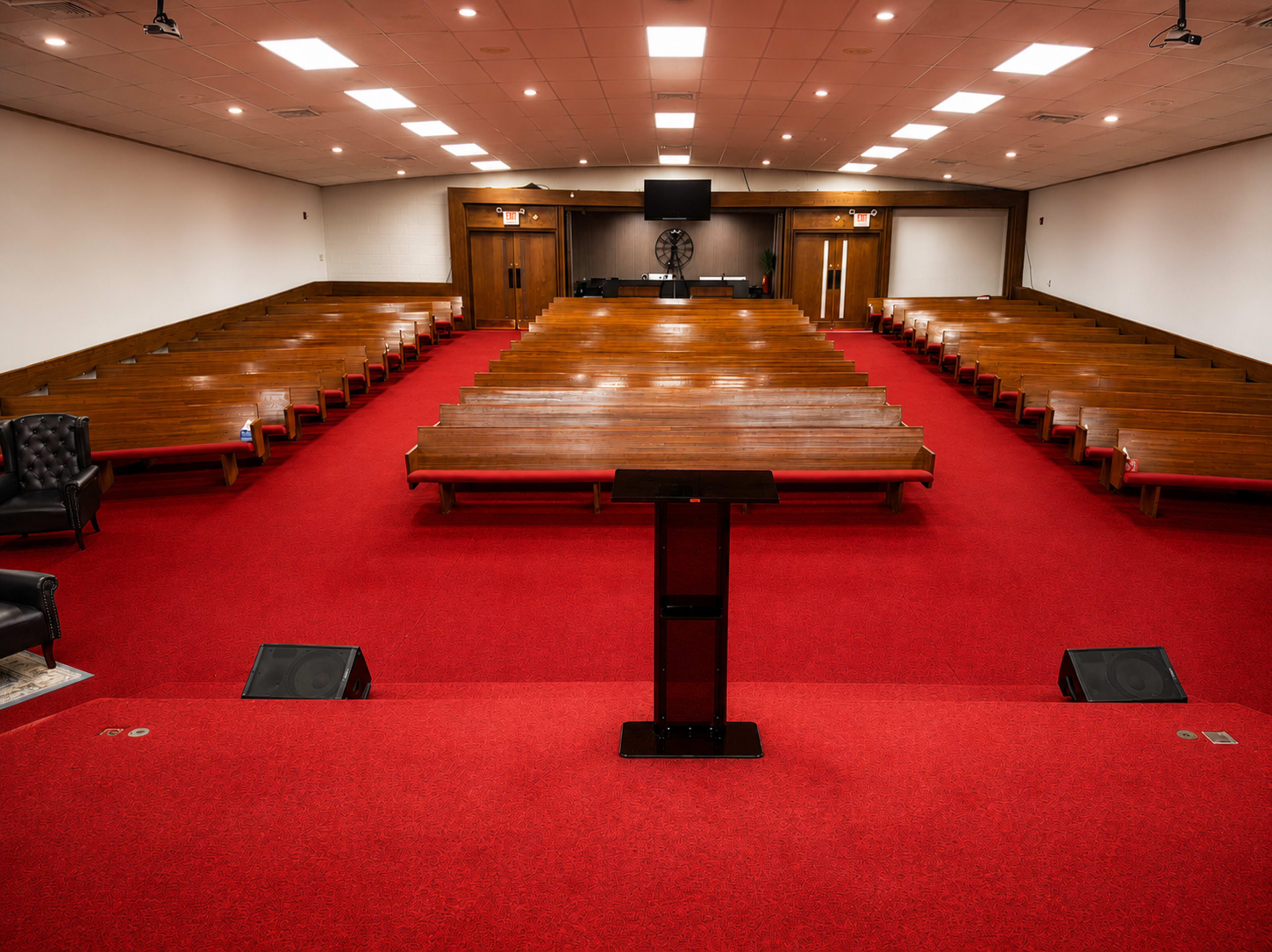 The image shows a spacious, empty church sanctuary with wooden pews arranged in rows on red carpeting, and a podium at the front facing the seating area.
