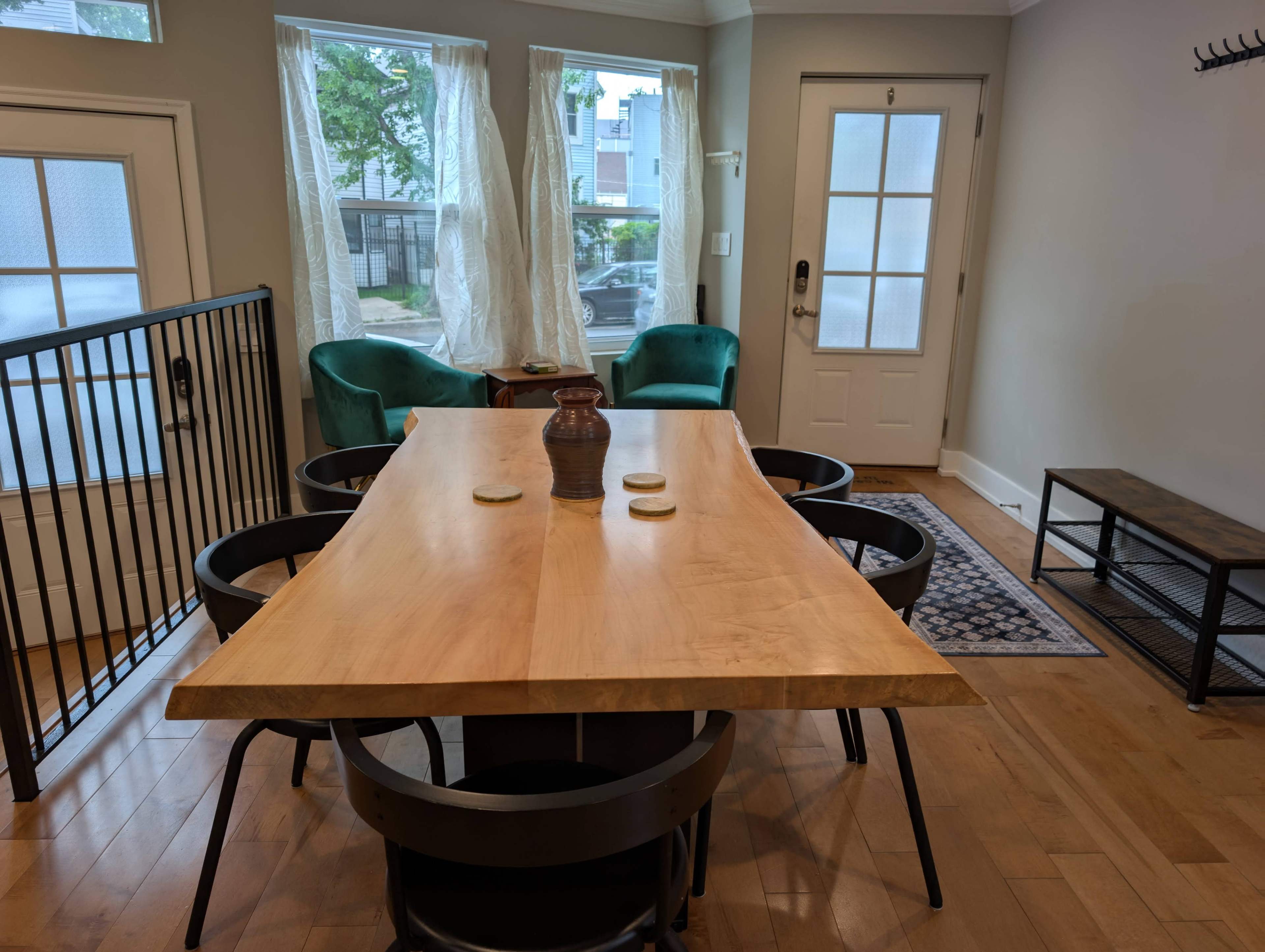 A dining area features a large wooden table surrounded by black chairs, with two green upholstered chairs in the background and large windows letting in natural light.