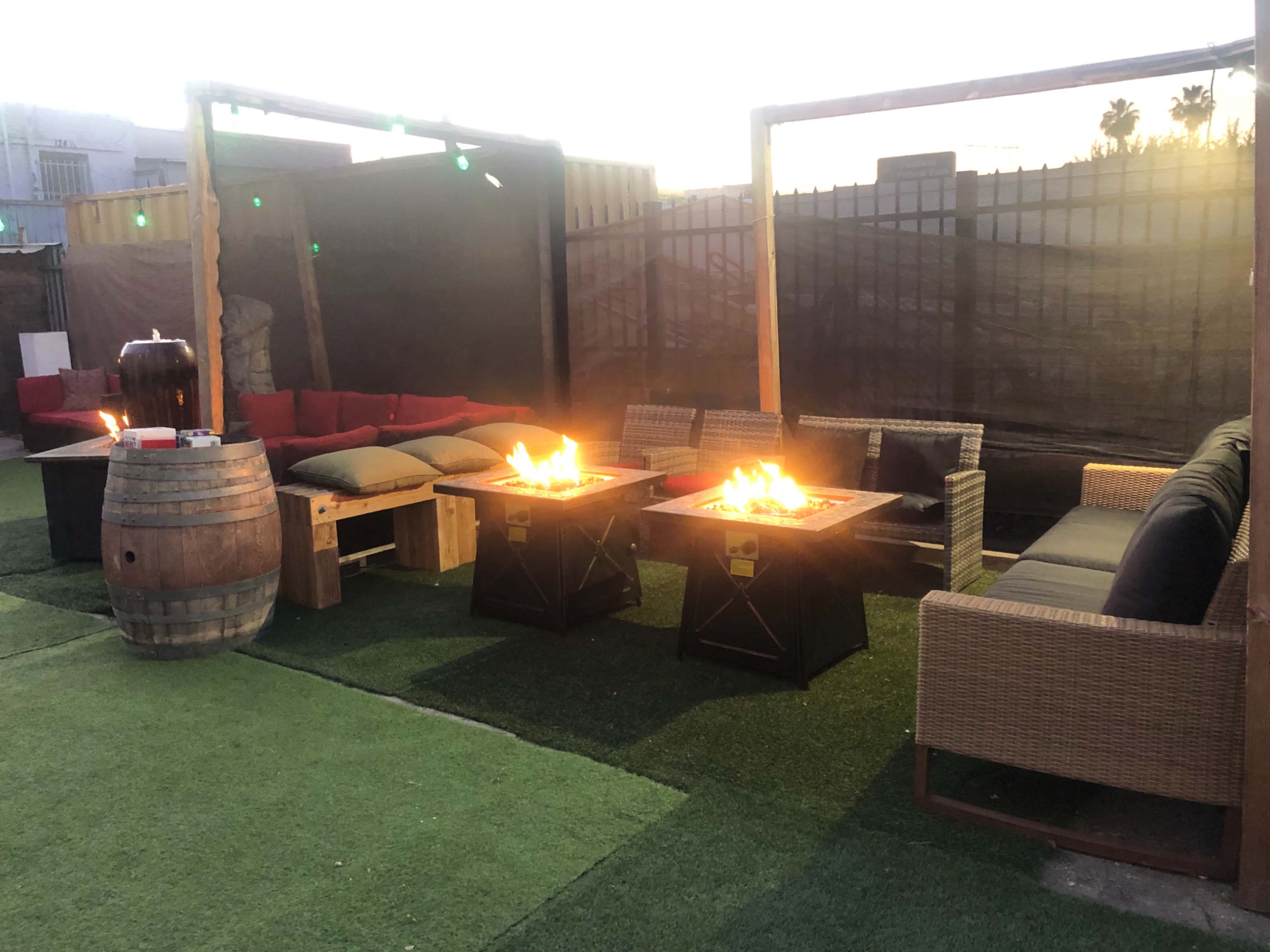The image shows an outdoor seating area with two fire pits surrounded by various couches and a wooden barrel.