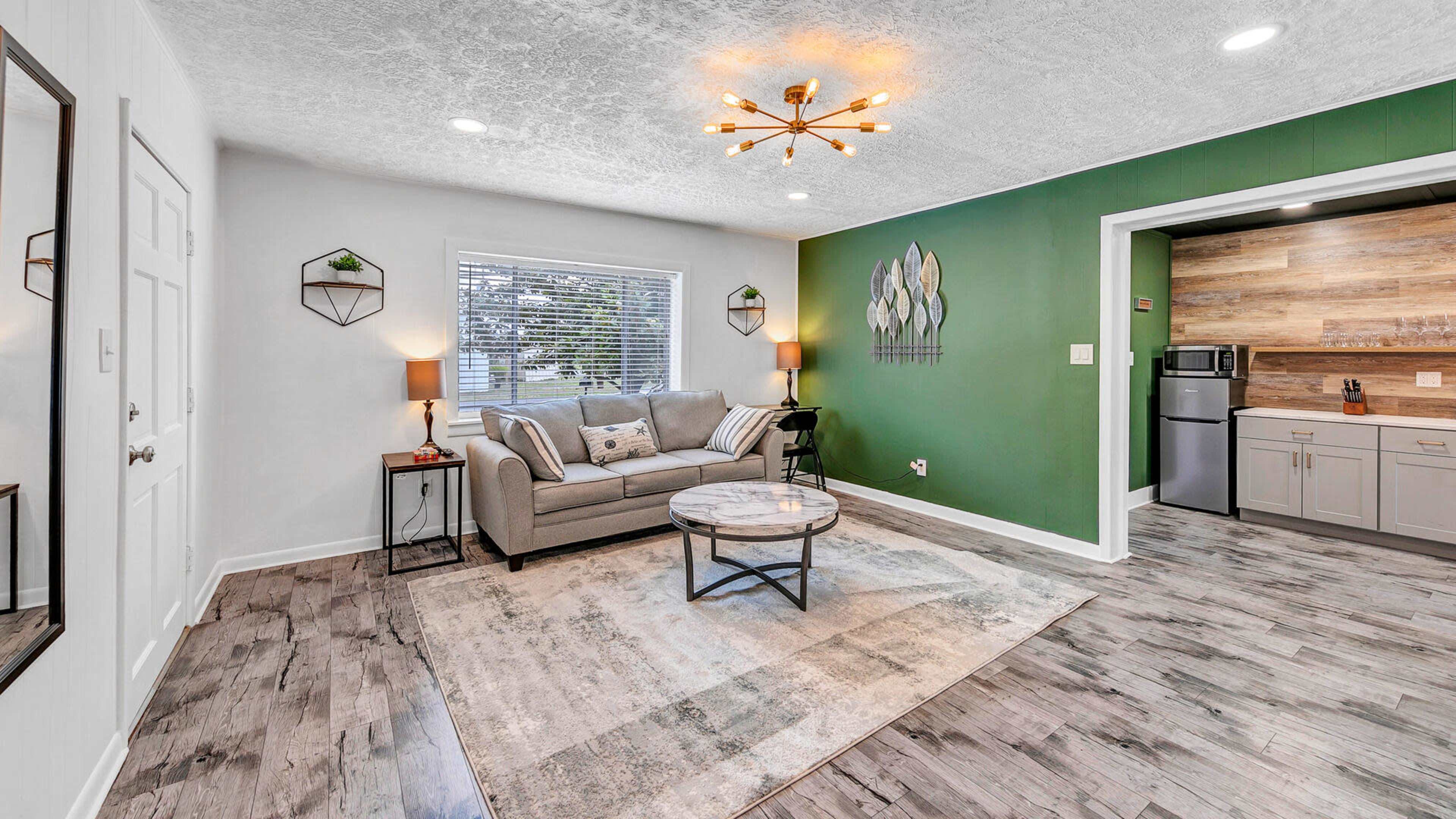 A modern living room with a gray sofa, a round coffee table, and green accent walls, leading to a kitchen area in the background.