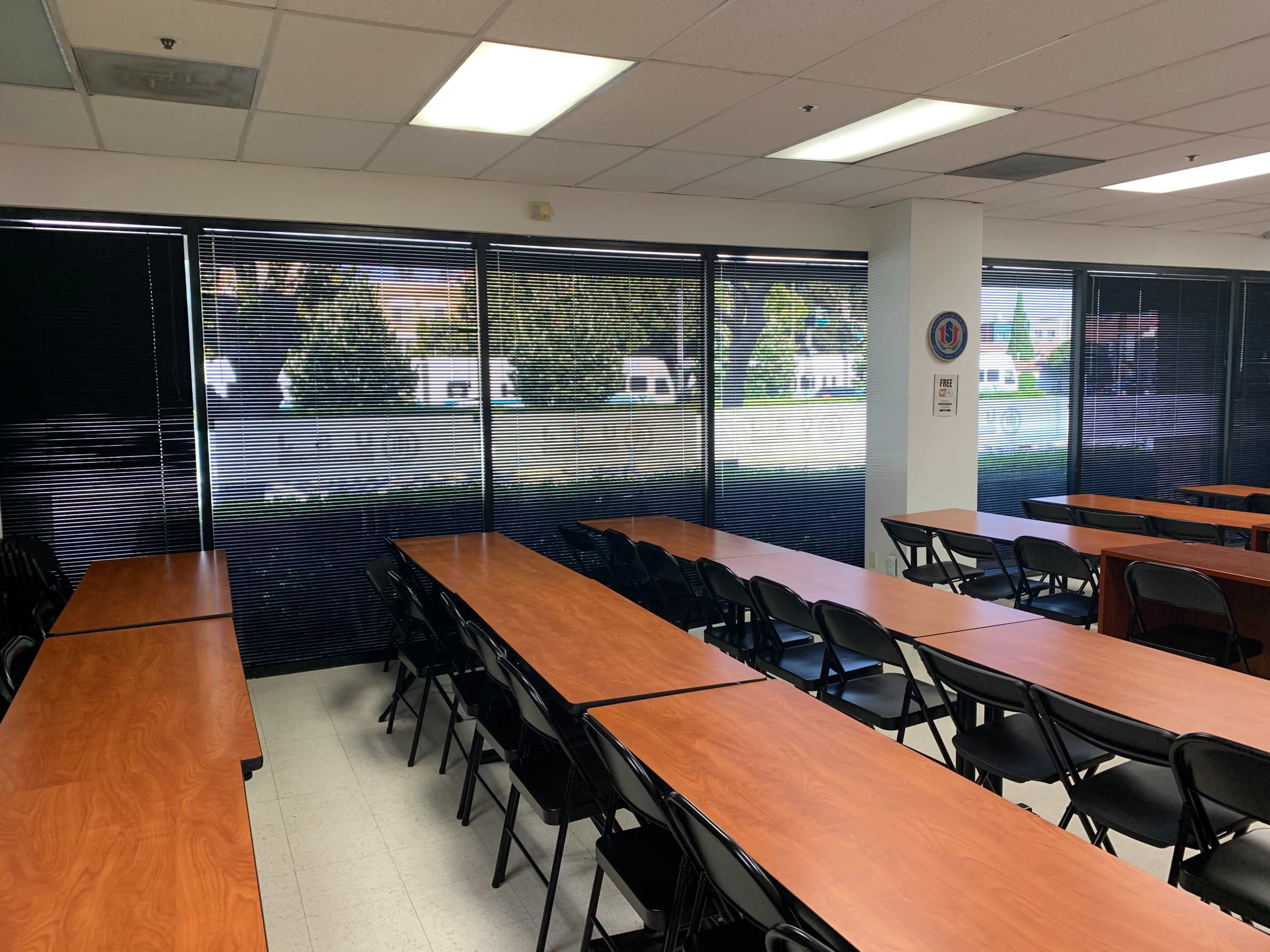 A classroom features long wooden tables arranged in rows with black chairs, and large windows showing a view of a landscaped outdoor area.