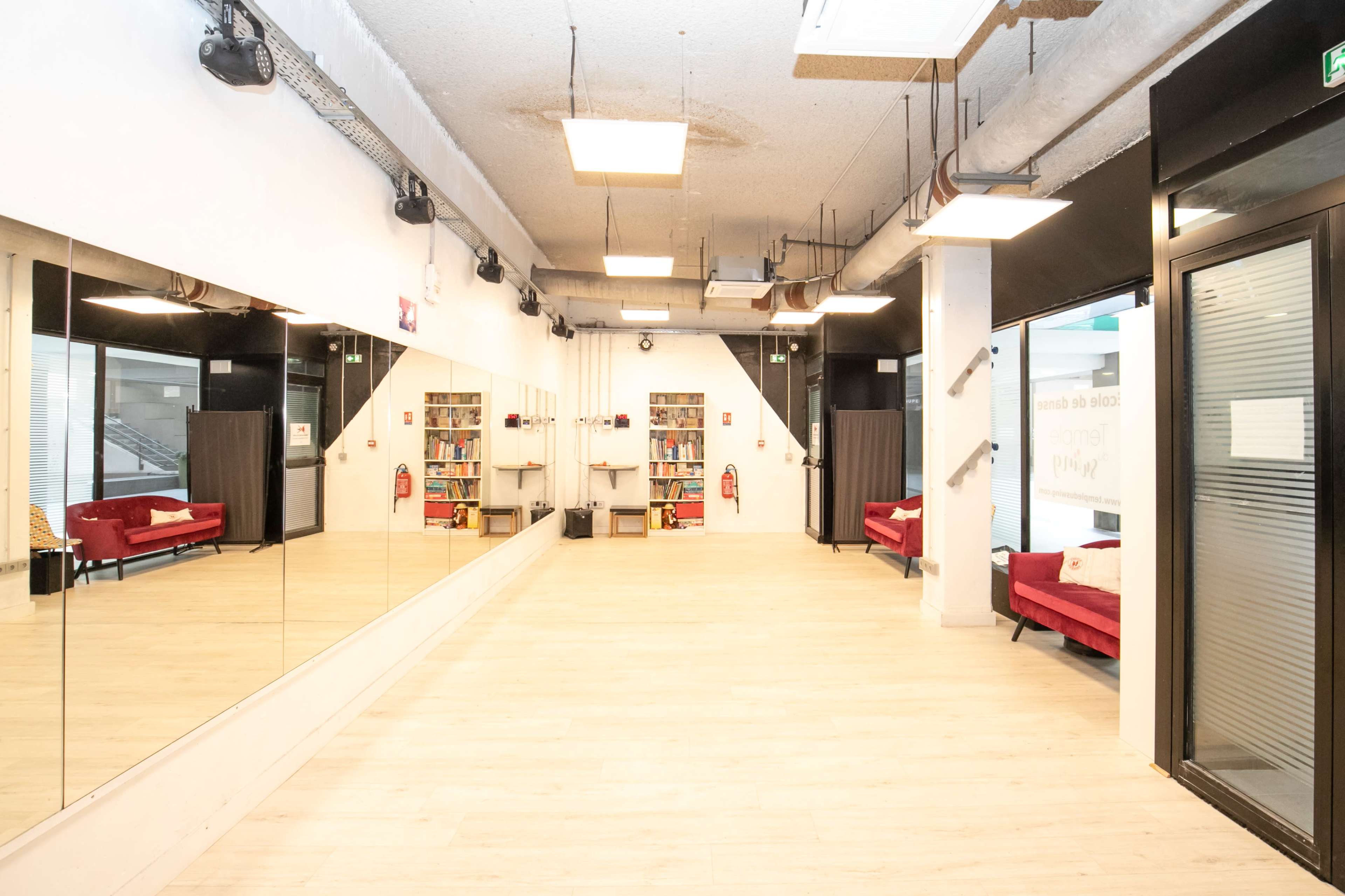A spacious room with wooden flooring, large mirrors on one wall, red sofas, and shelves filled with books.