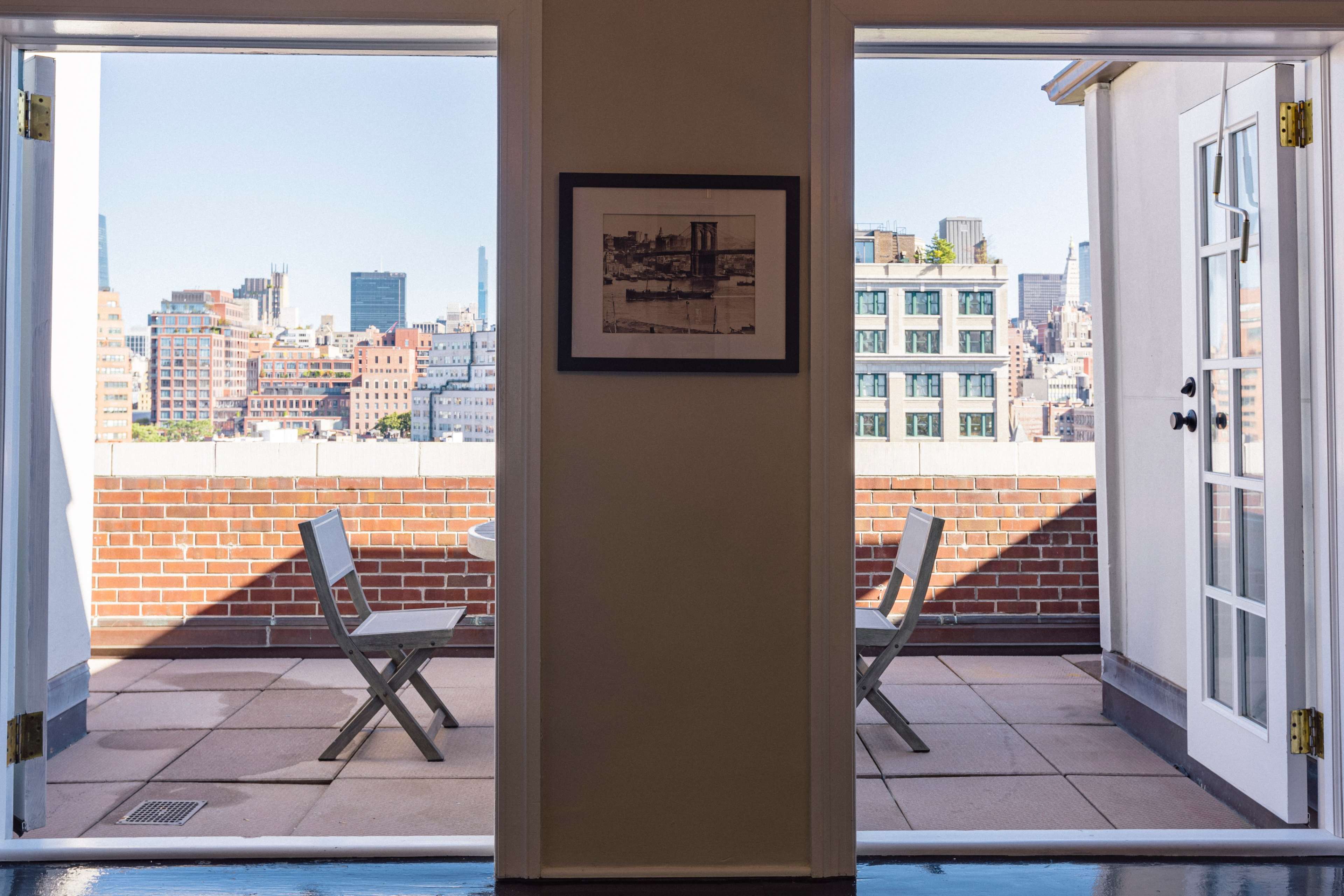 A balcony with two chairs and a small table overlooks a city skyline through open double doors.