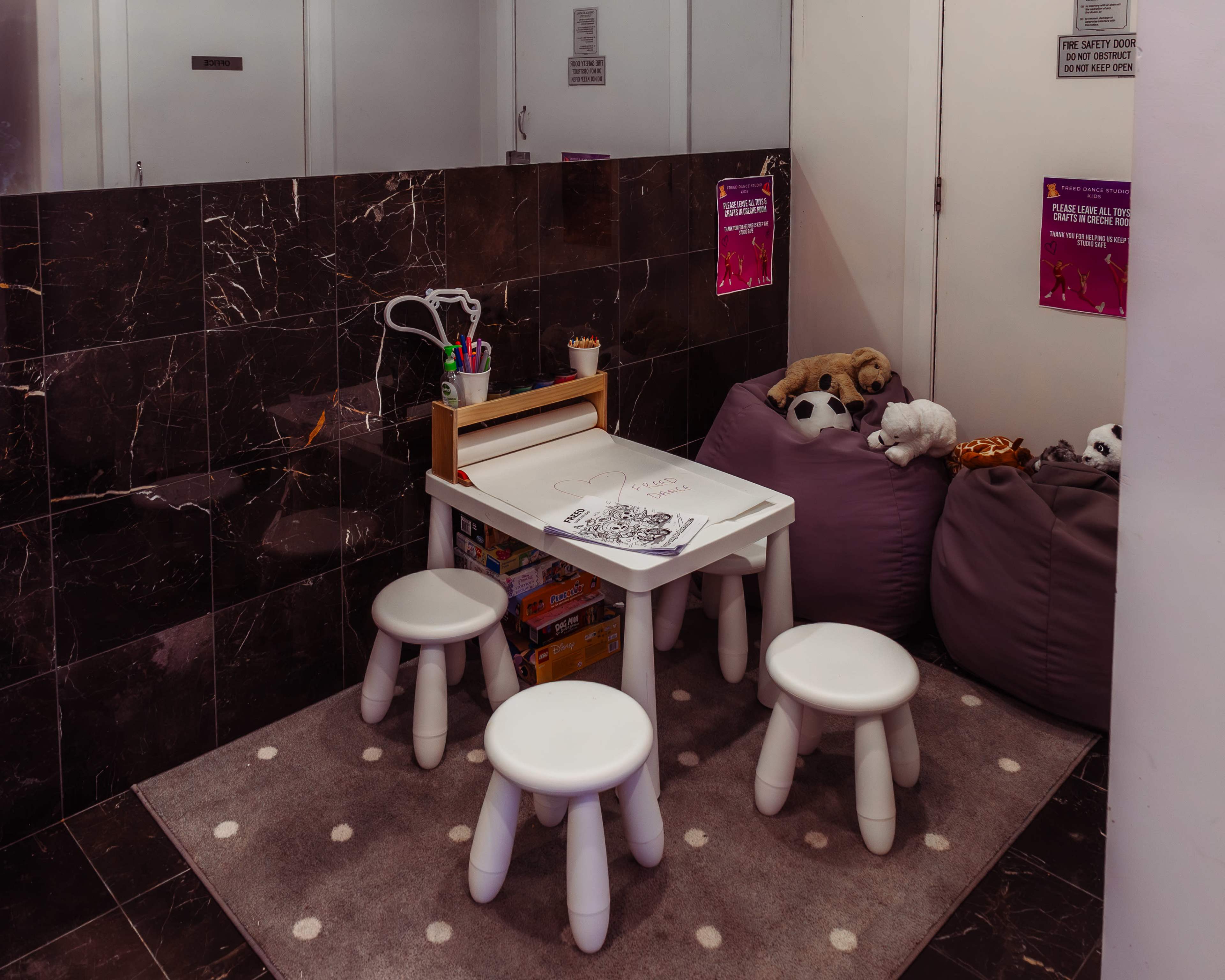 The image shows a small play area with a white table and four stools surrounded by dark marble walls, featuring stuffed animals and art supplies.