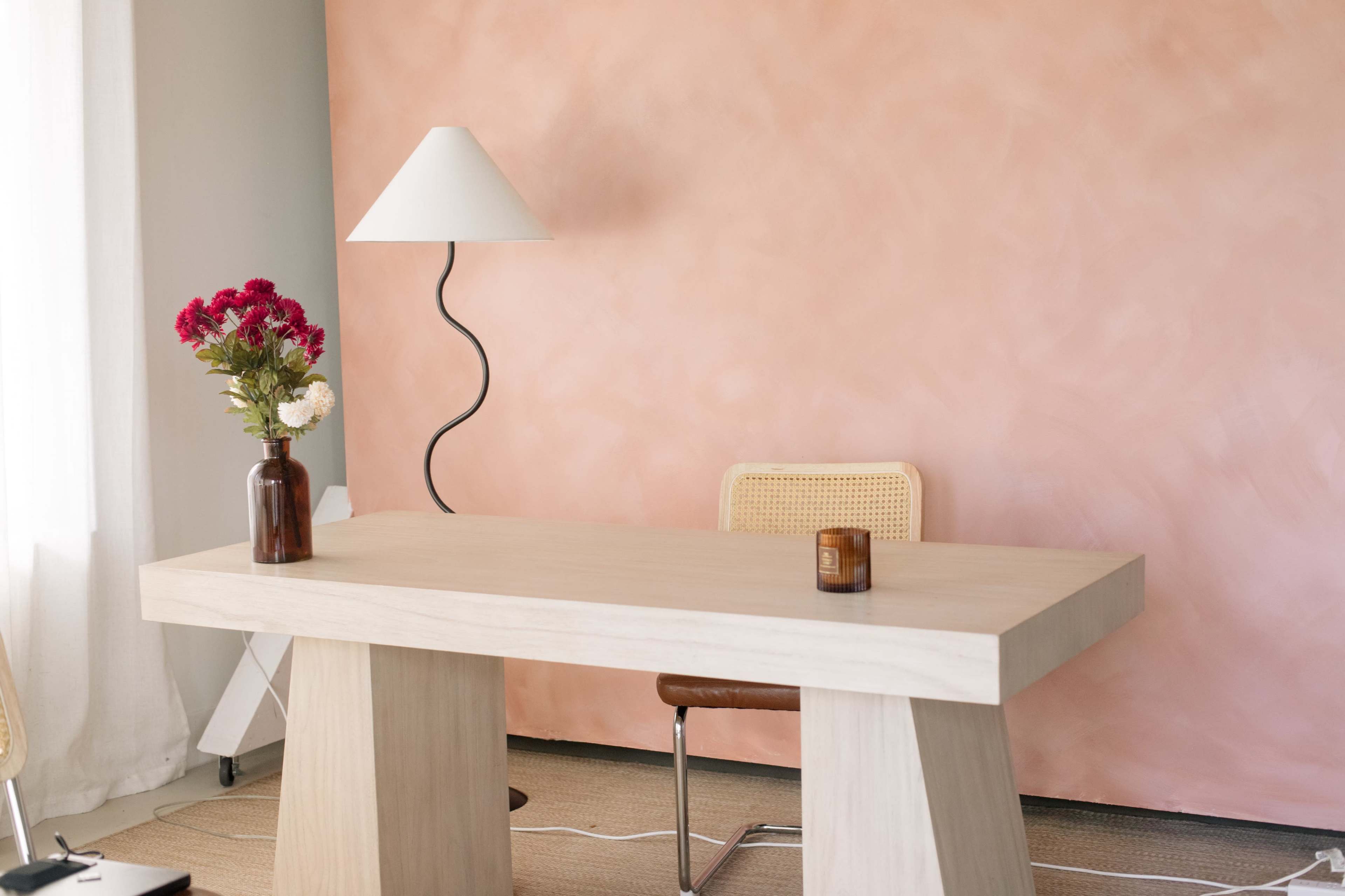 A simple workspace features a wooden desk, a chair, a lamp, and a vase of flowers against a pink wall.