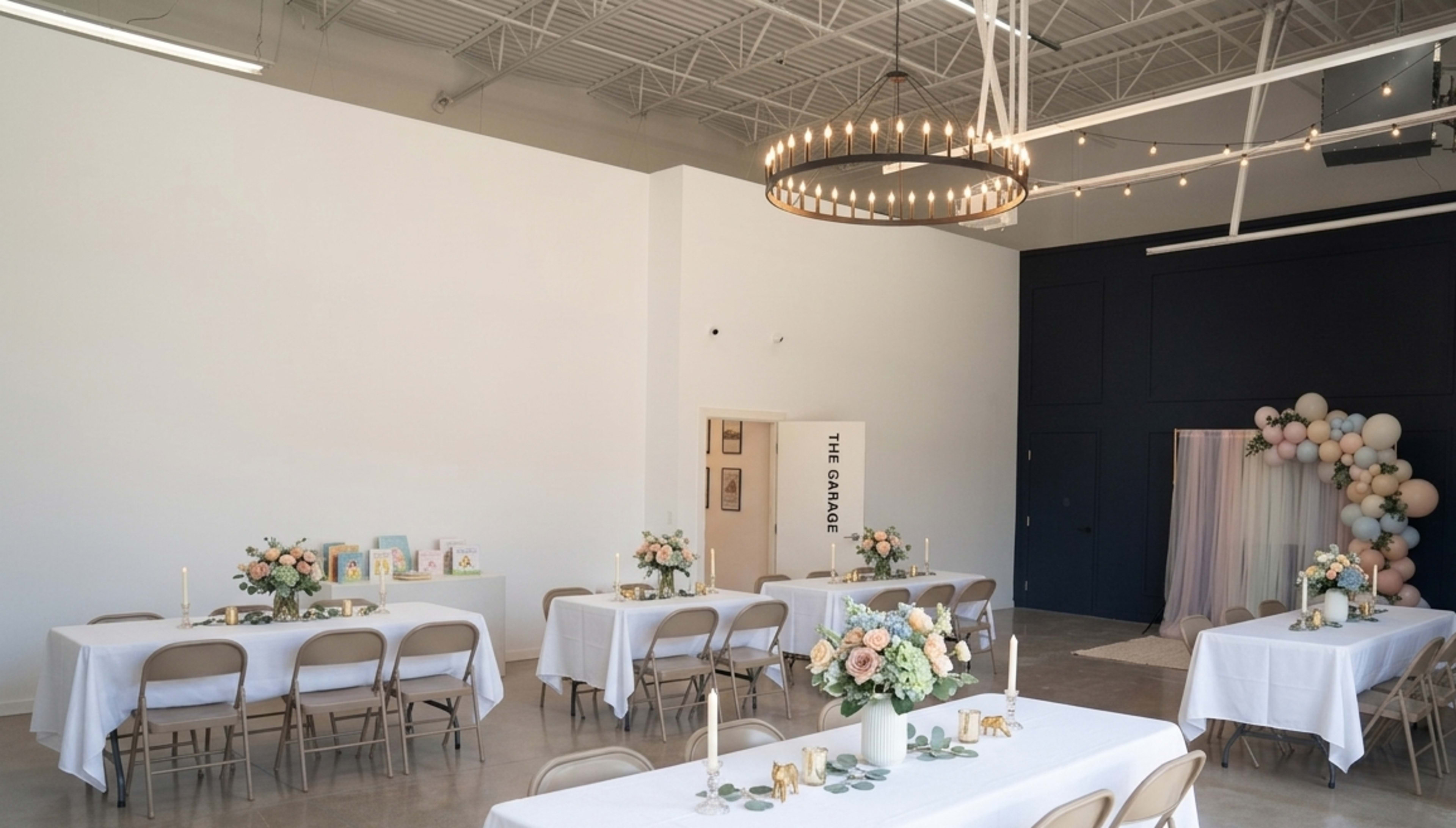 The image shows a spacious event space with tables set for an occasion, featuring white tablecloths, floral centerpieces, candles, and a decorative background with balloons.
