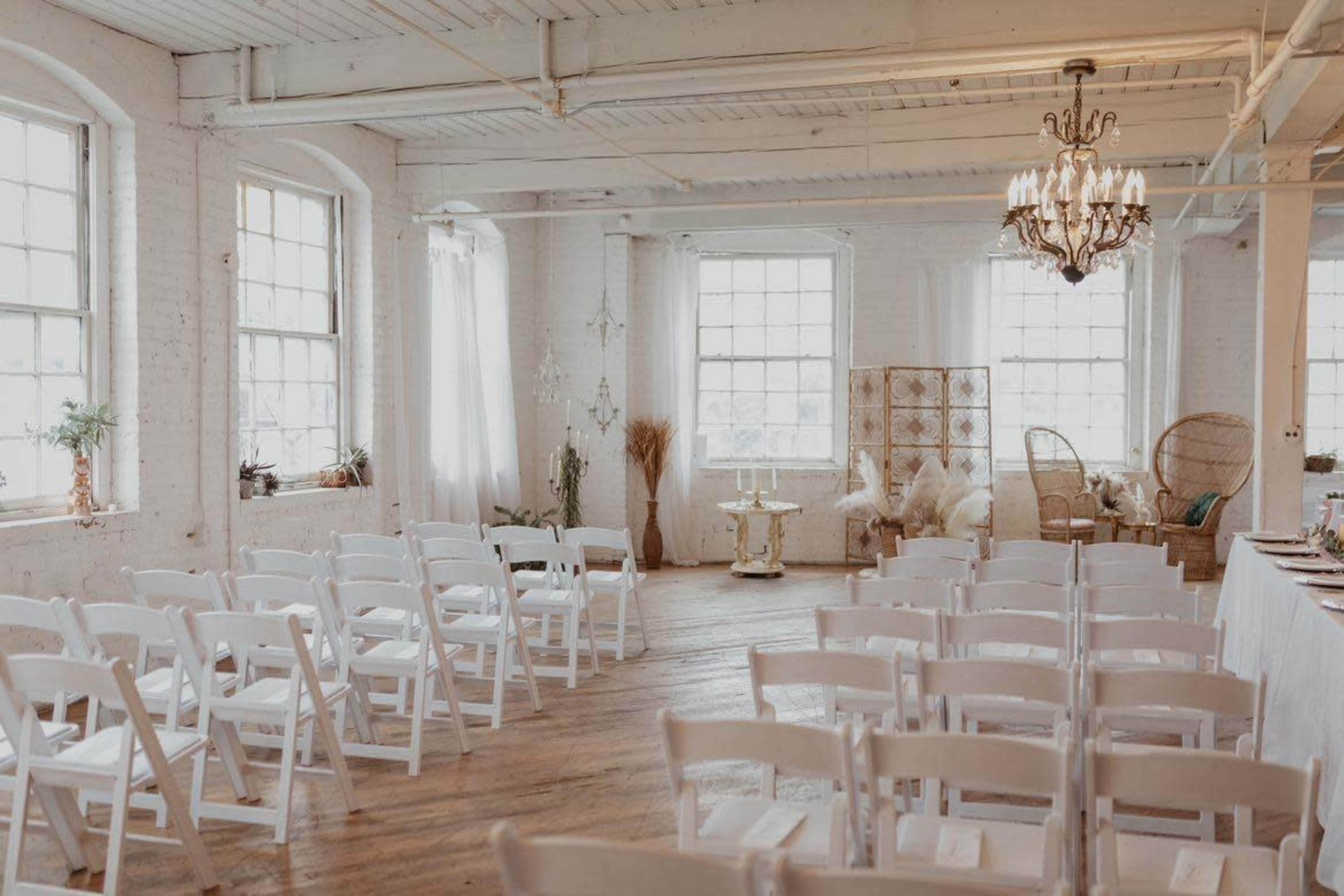 The image shows a bright, spacious event venue with rows of white chairs arranged for an occasion, a chandelier hanging from the ceiling, and various decorative elements along the walls.