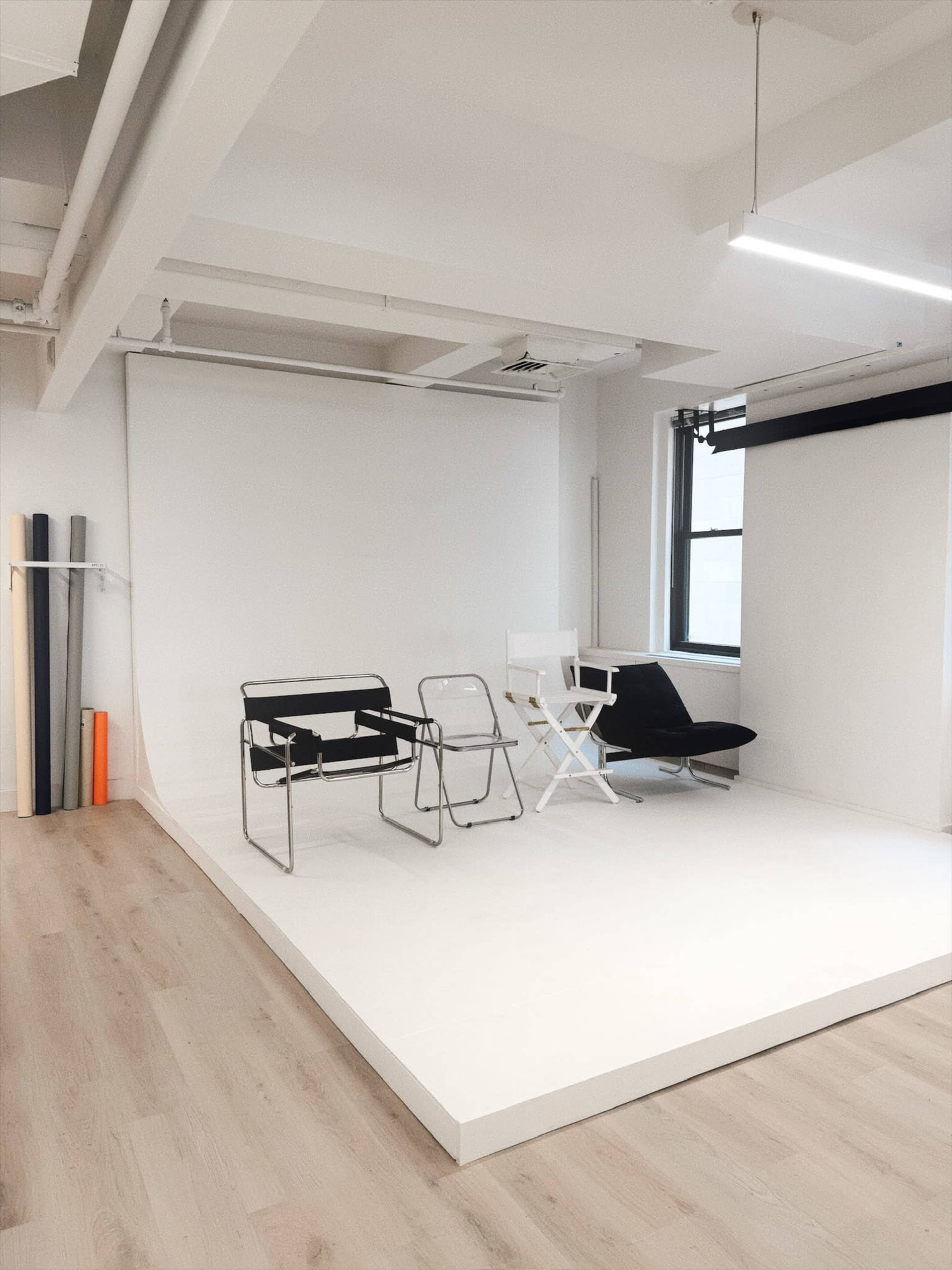 The image shows a minimalist studio space with a white backdrop and three chairs on a light wooden floor.