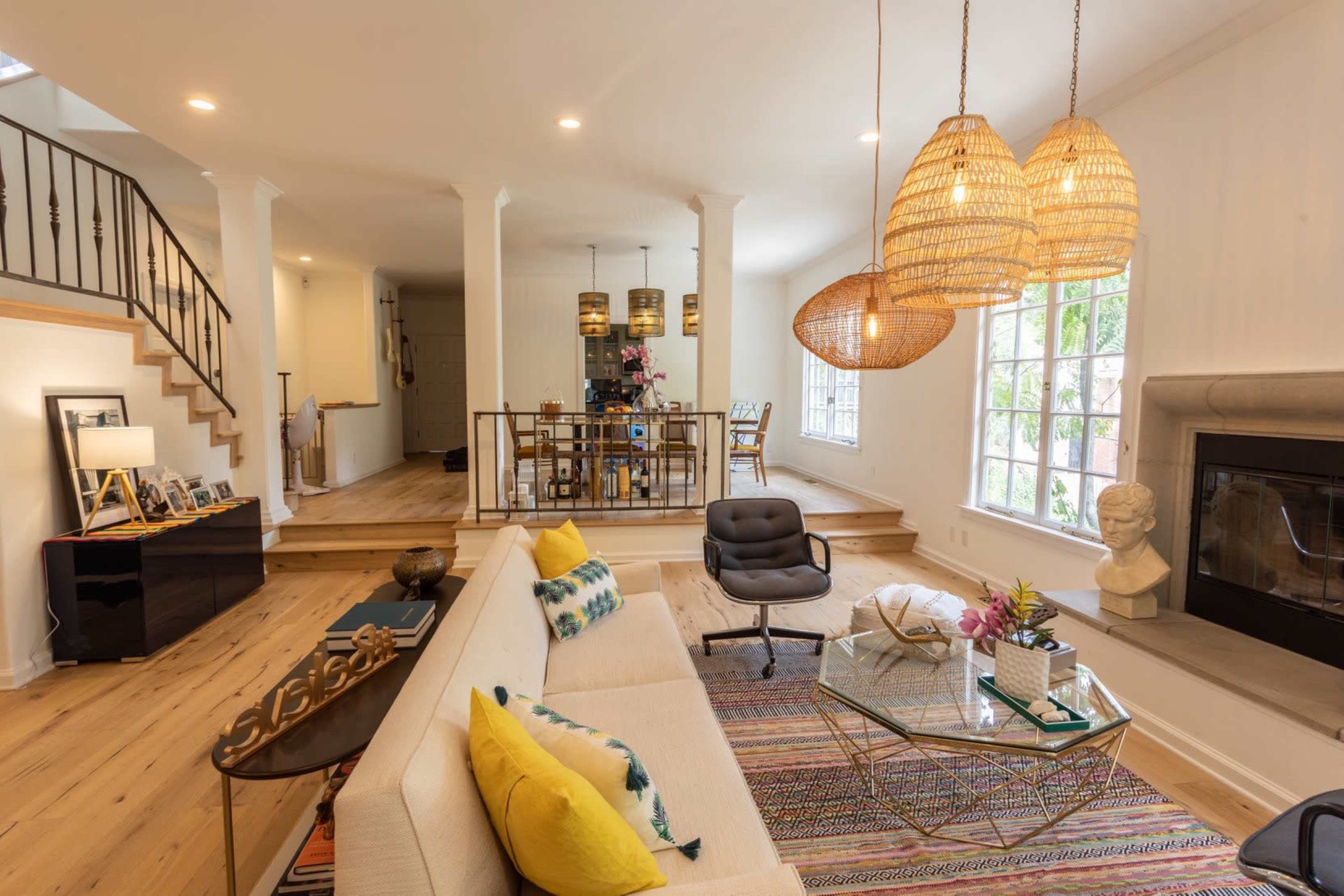 A modern living room features a cream-colored sofa with yellow cushions, a glass coffee table, and large woven pendant lights, with a dining area and staircase visible in the background.