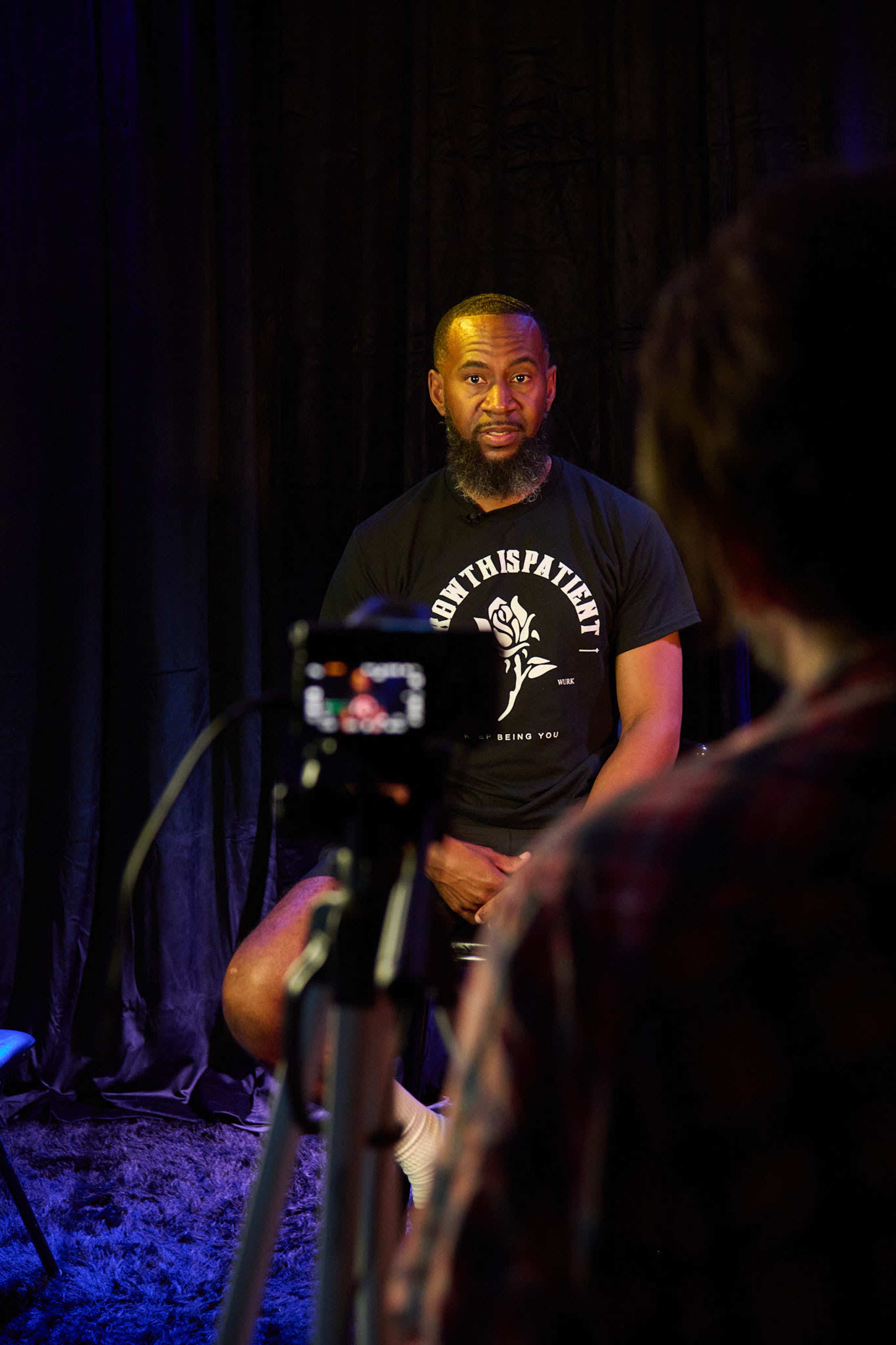 A man wearing a black t-shirt sits in front of a camera, while another person in a checkered shirt is partially visible in the foreground.