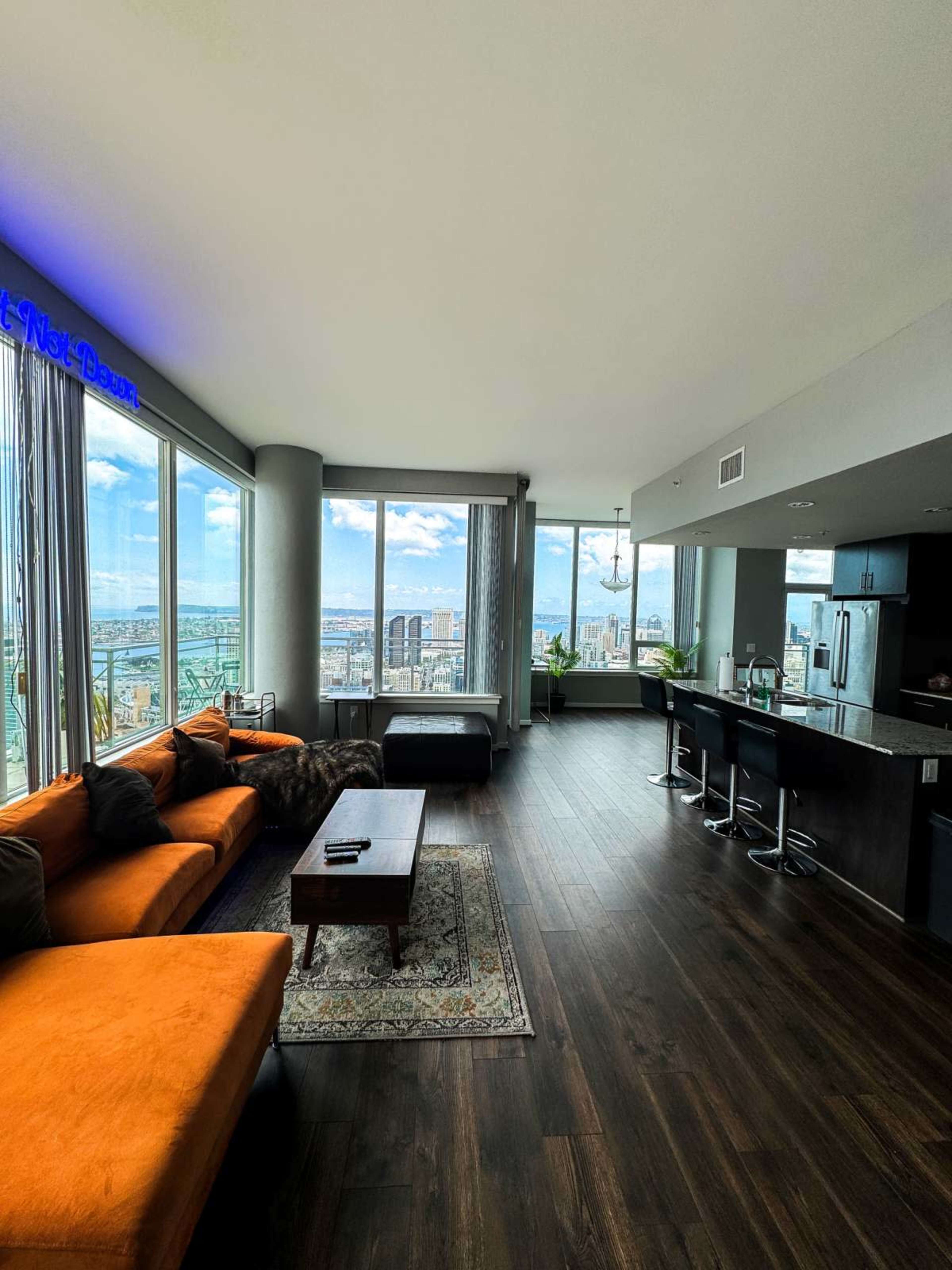 A modern living room with a large orange sofa, a wooden coffee table, and a kitchen area, all overlooking a city skyline through large windows.