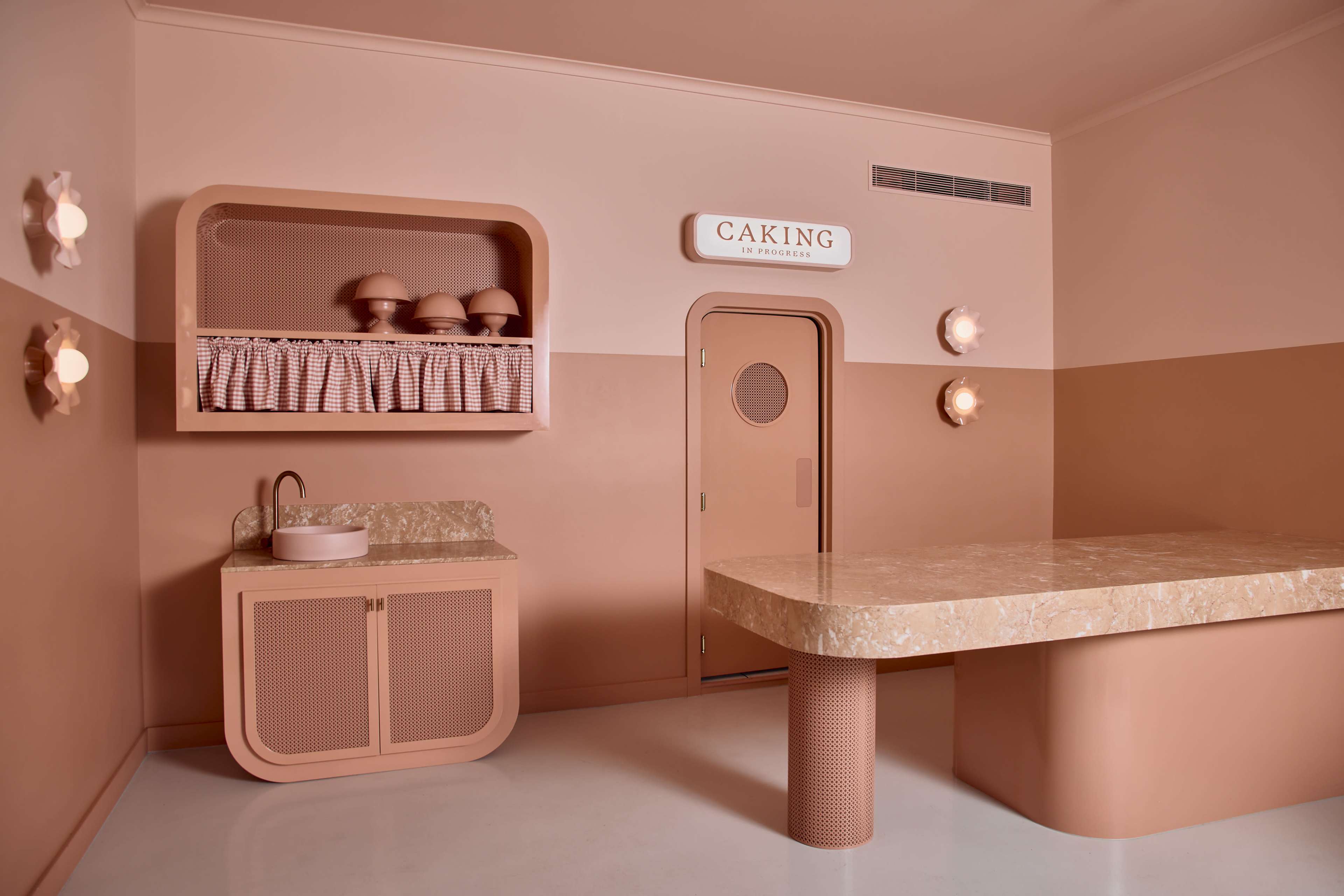 The image shows a minimalist interior space with beige and peach walls, featuring a large marble-topped table, a washing area, and a door labeled "CAKING."