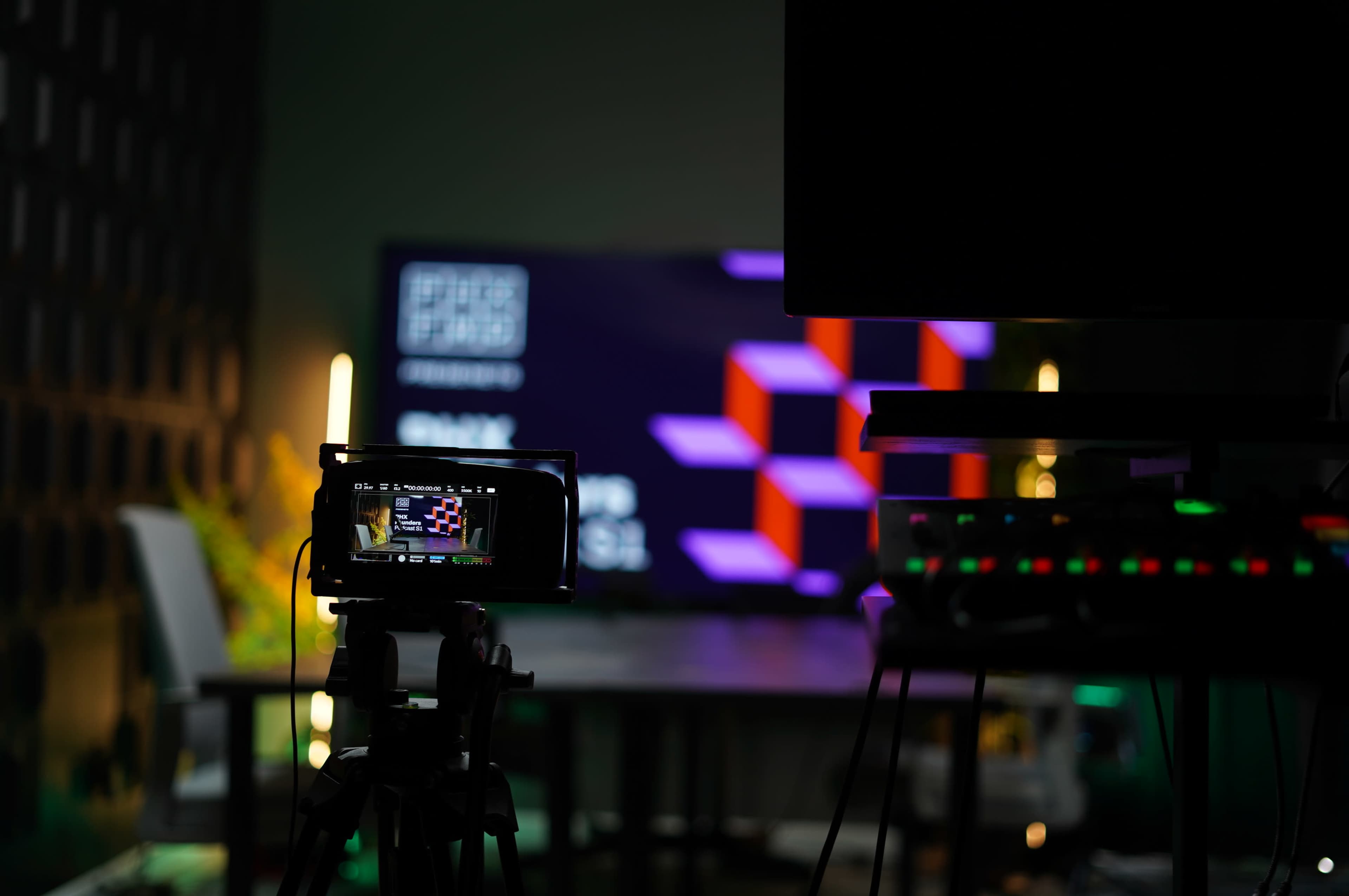 A camera on a tripod focuses on a large television screen displaying colorful graphics in a dimly lit studio setting.