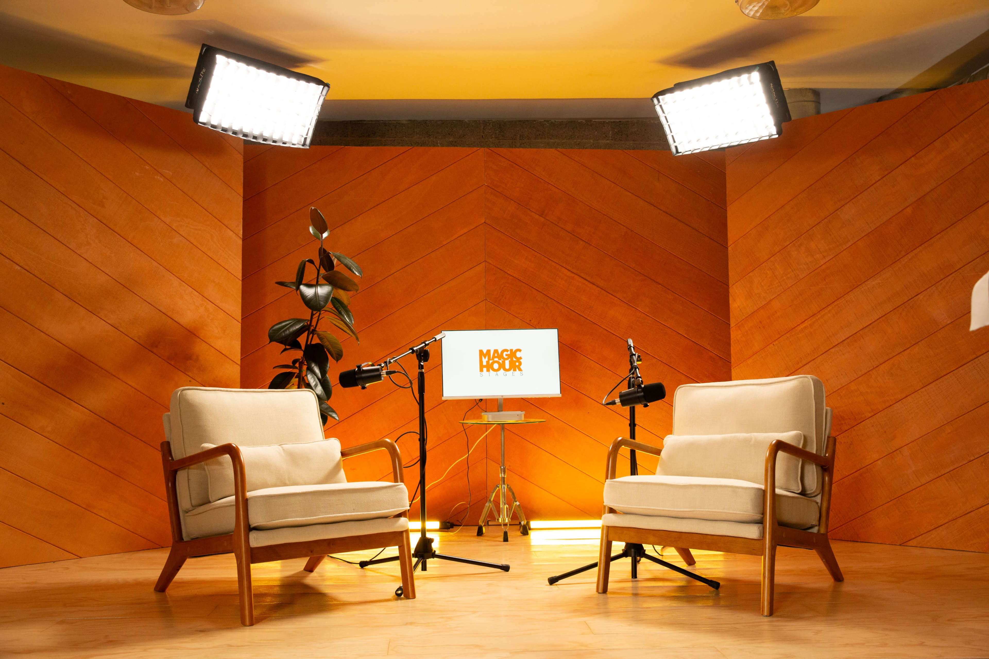 The image shows a cozy interview setup featuring two cream-colored chairs, a small table with a monitor displaying "MAGIC HOUR," and soft lighting against a wooden background.
