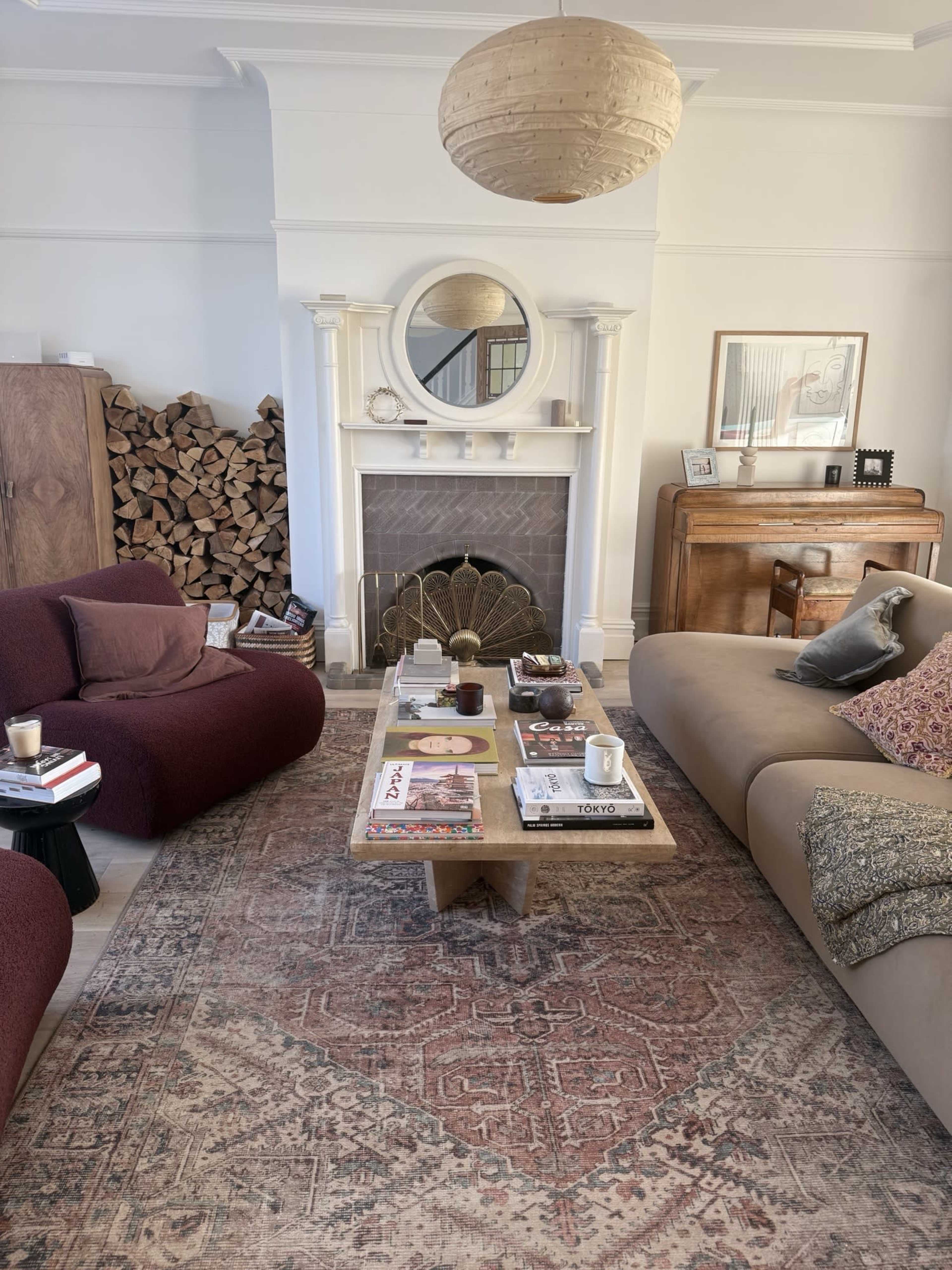 The living room features a fireplace with a round mirror above it, a wooden coffee table surrounded by a burgundy and a beige sofa, and neatly stacked firewood in the corner.