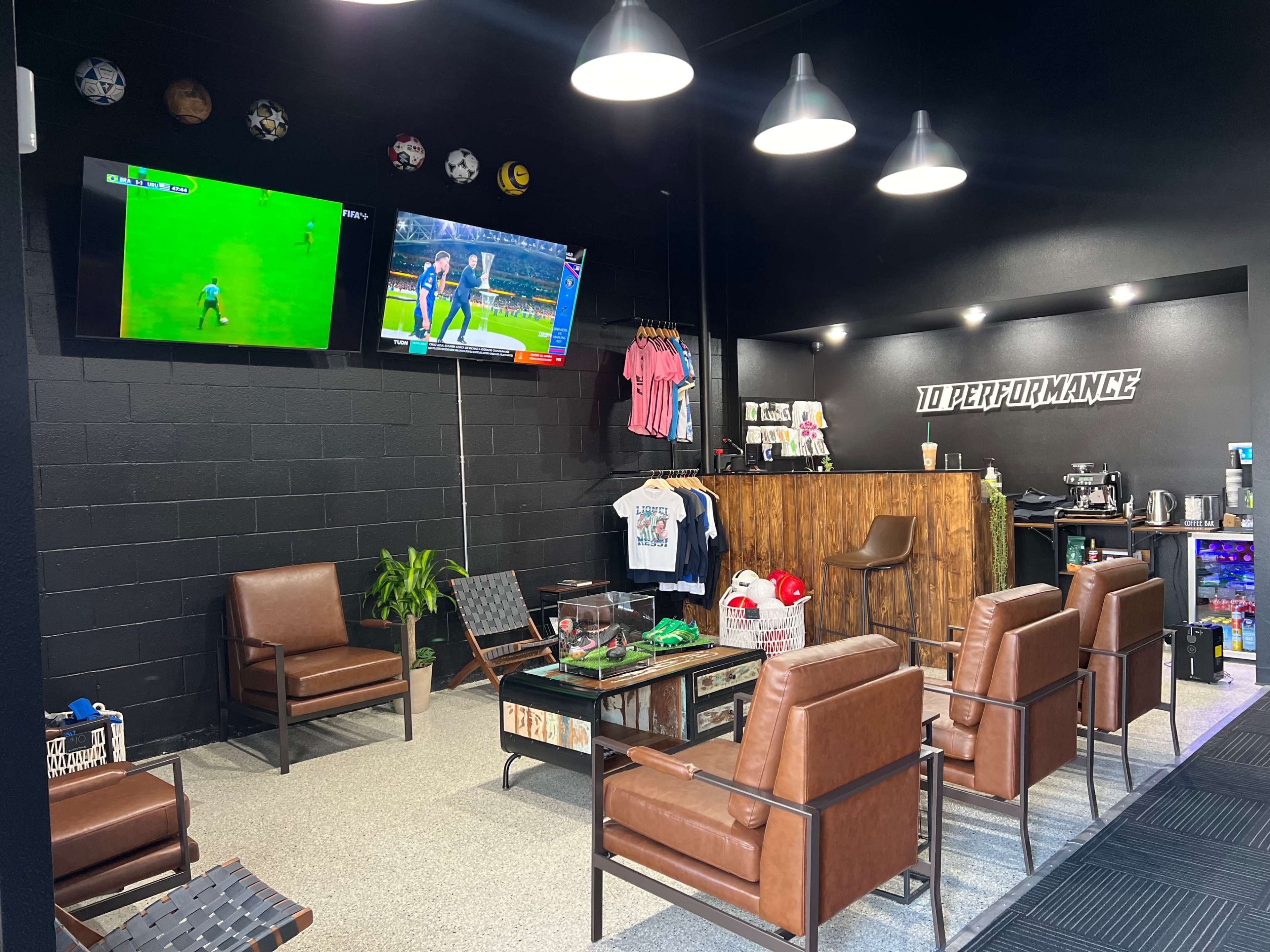 A modern lounge area with brown leather chairs, two televisions displaying sports, a wooden bar counter, and merchandise hanging on the wall.