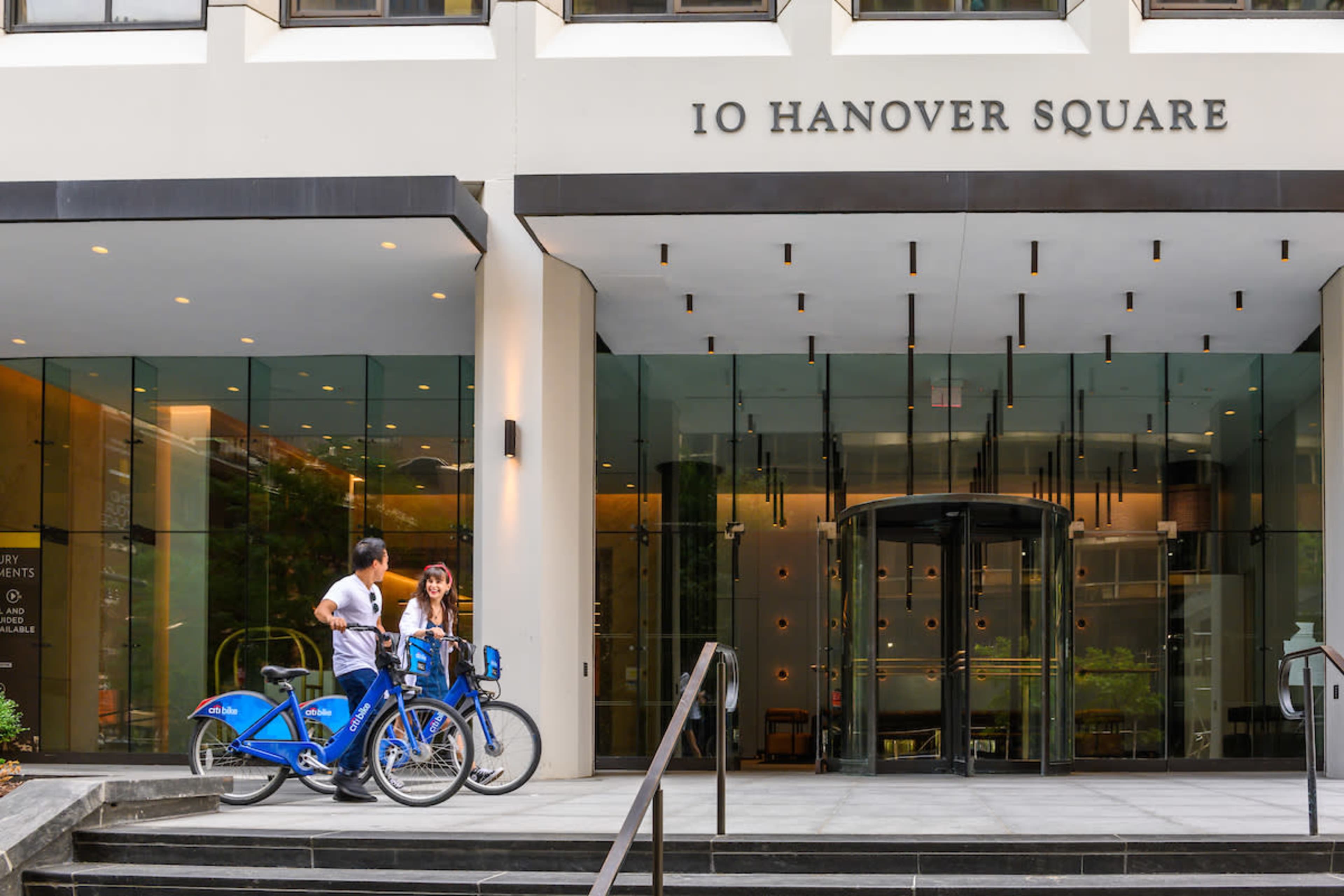 Two people are standing next to blue bicycles in front of a modern building with the sign "10 Hanover Square."