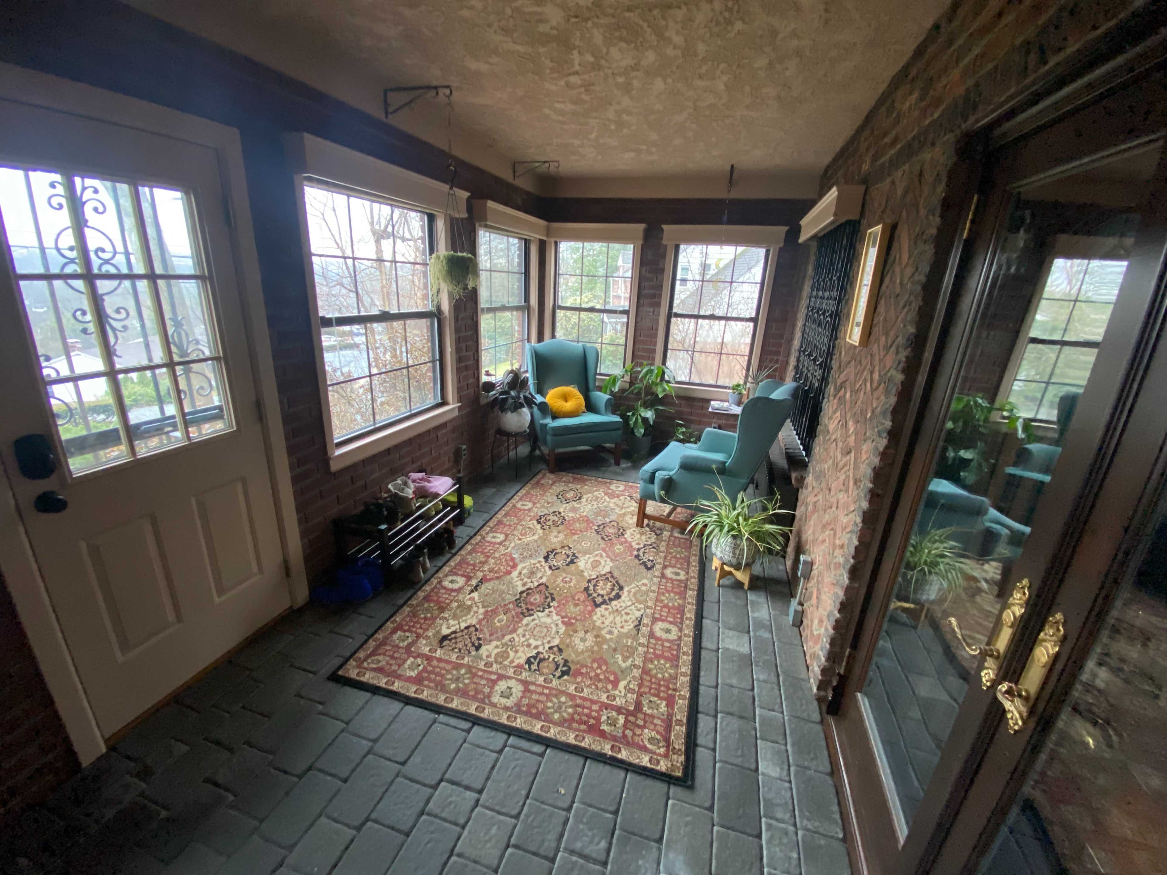 A sunroom with two teal armchairs, a patterned rug, potted plants, and large windows overlooking a natural landscape.
