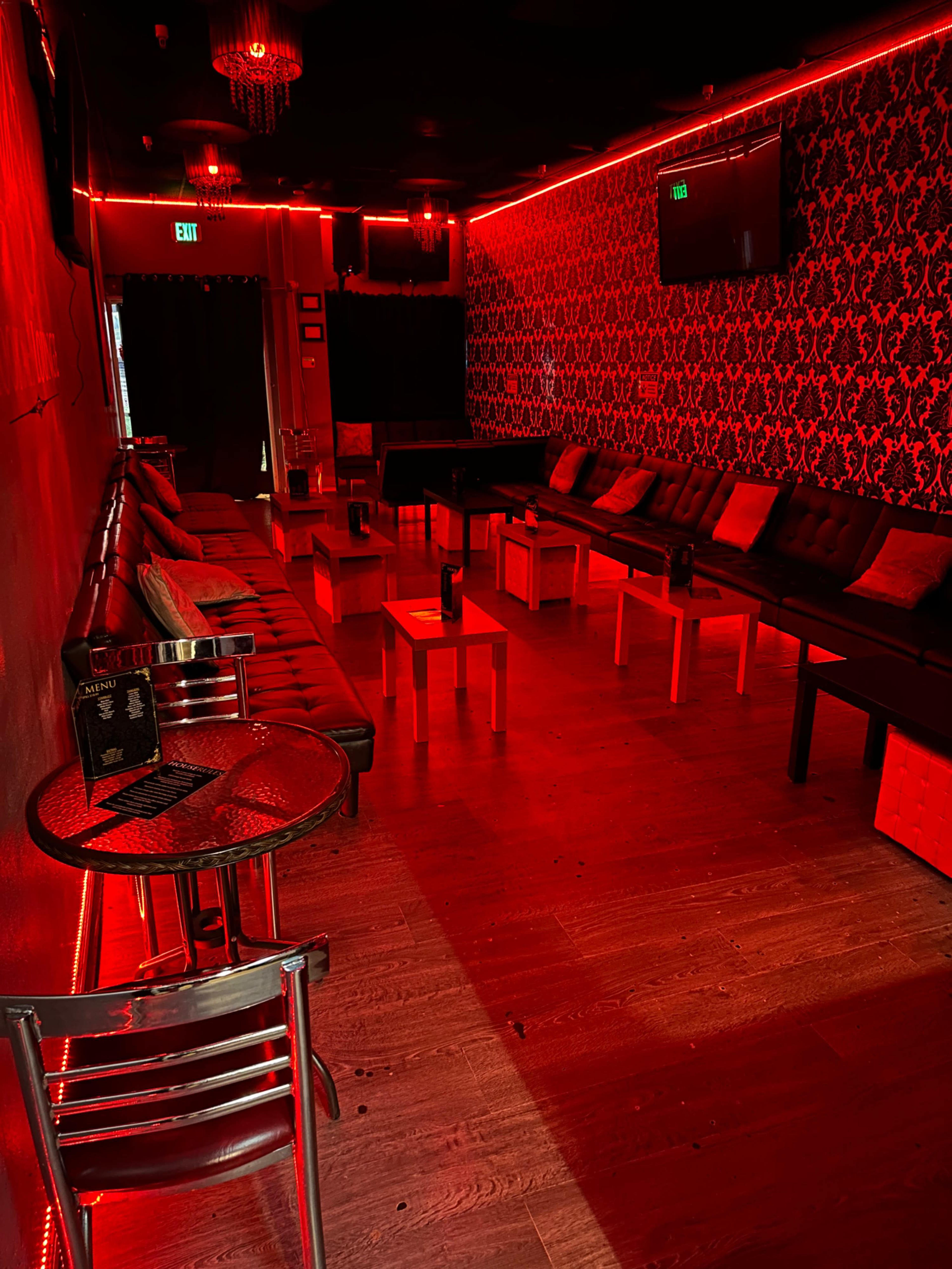 The image shows a dimly lit lounge with red ambient lighting, featuring black couches, small tables, and a patterned wallpaper.