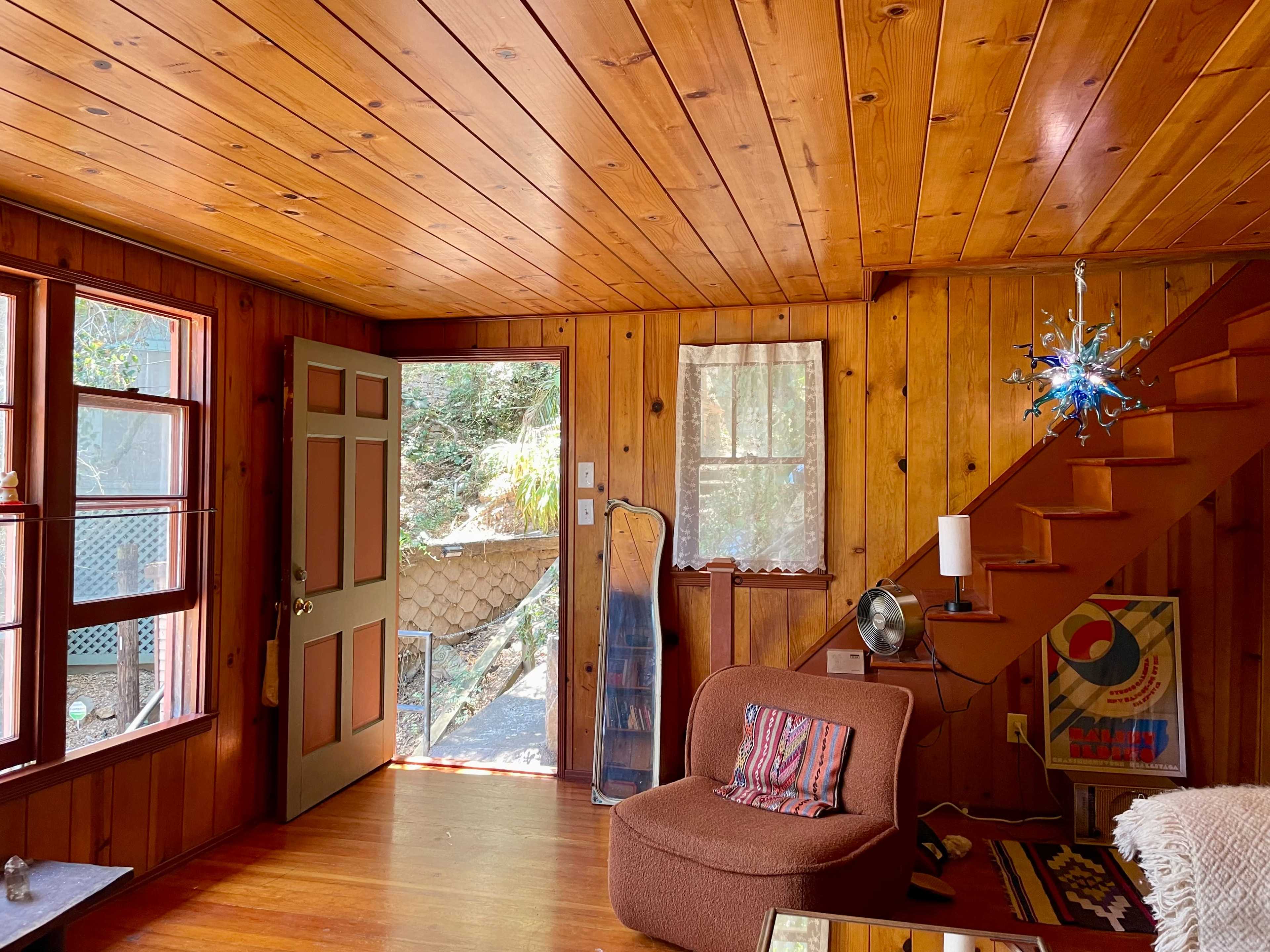 The image depicts a cozy wooden-paneled living room with a staircase, an open door leading outside, and a small seating area.