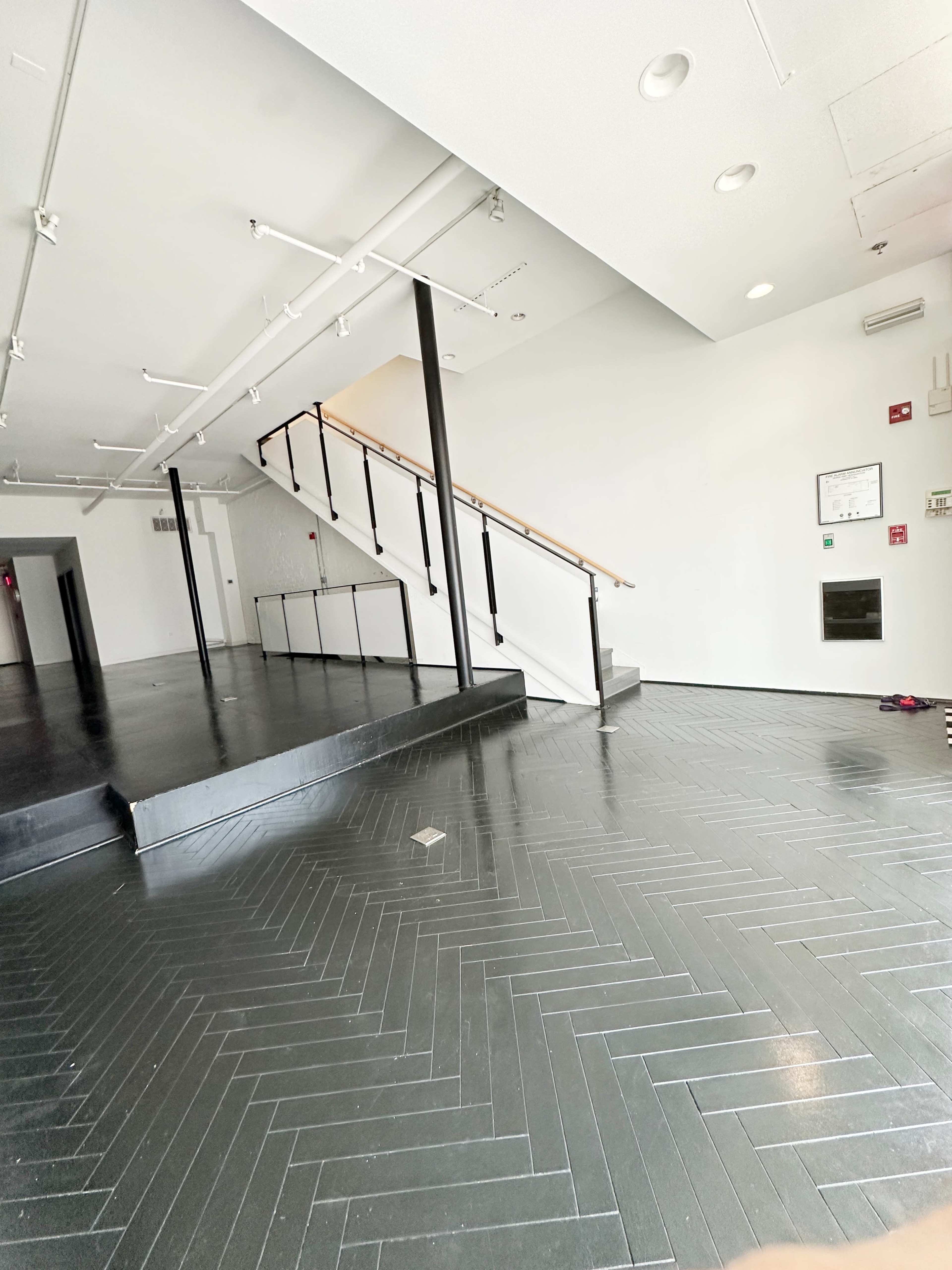 The image shows an empty, spacious interior with a staircase leading to an upper level and a polished herringbone-patterned floor.