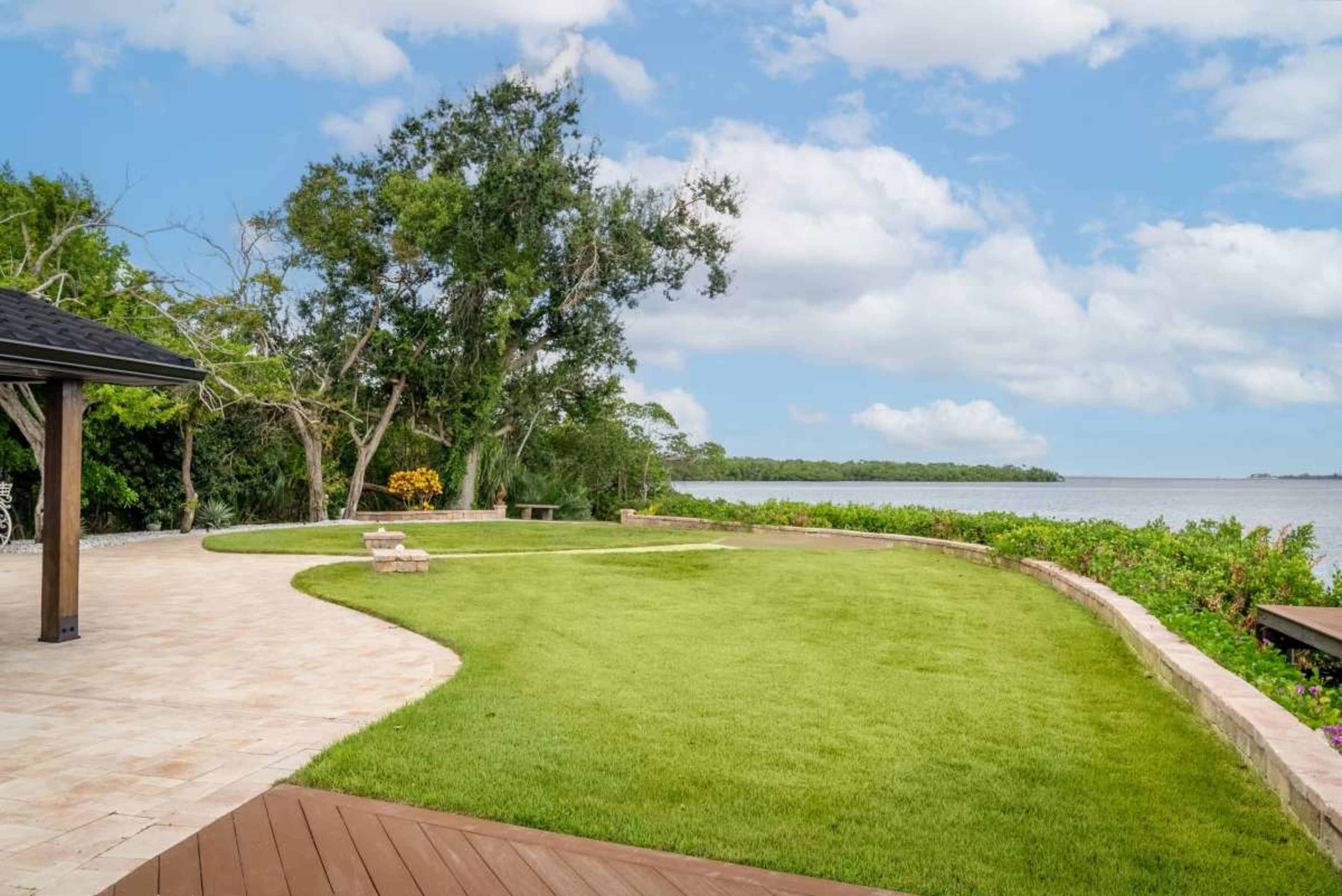 The image shows a landscaped outdoor area with a lush green lawn, stone pathways, and a view of the water surrounded by trees.