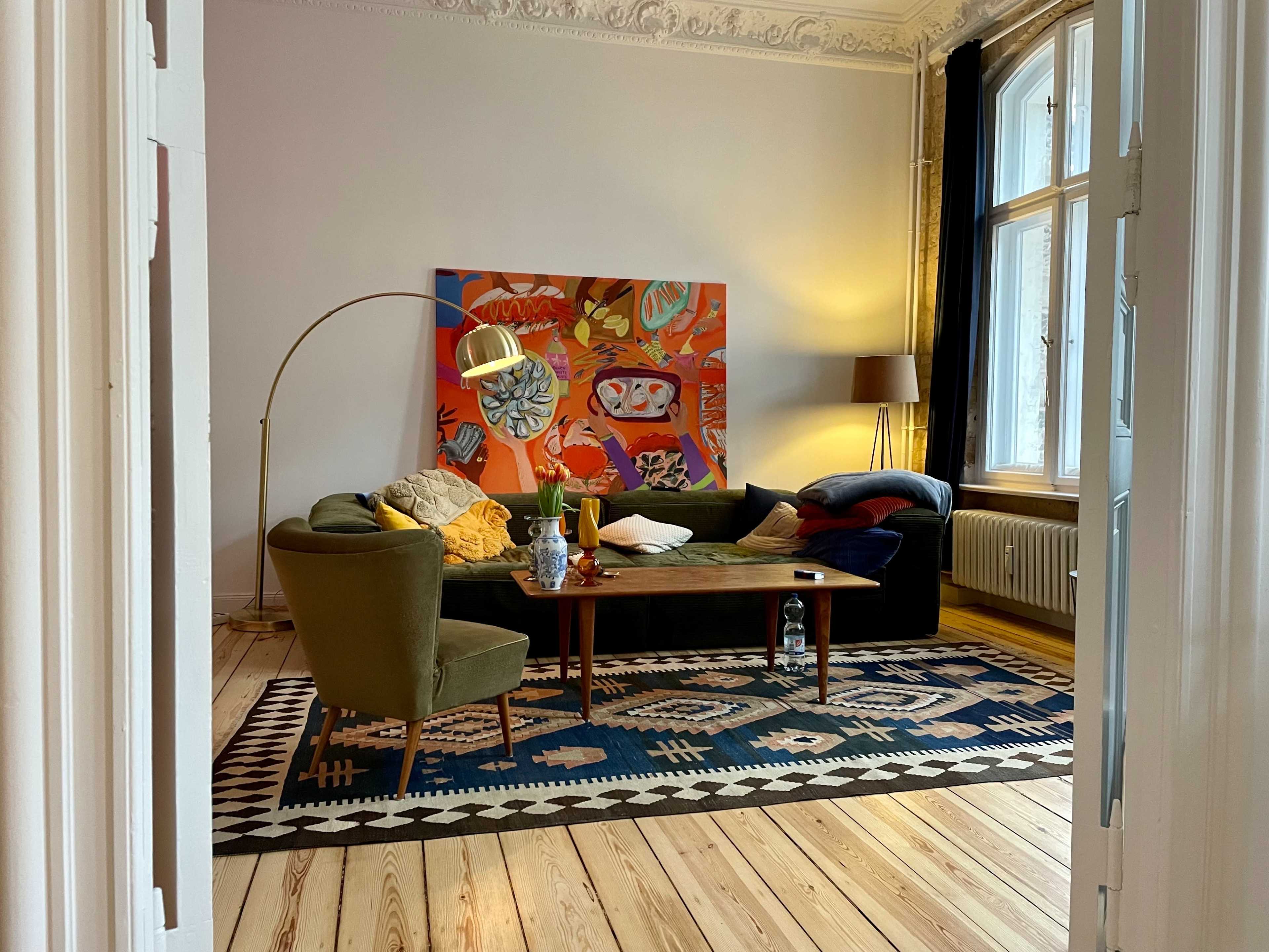 A cozy living room features a dark green sectional sofa, a wooden coffee table, a large colorful artwork on the wall, and a floor lamp beside a window.