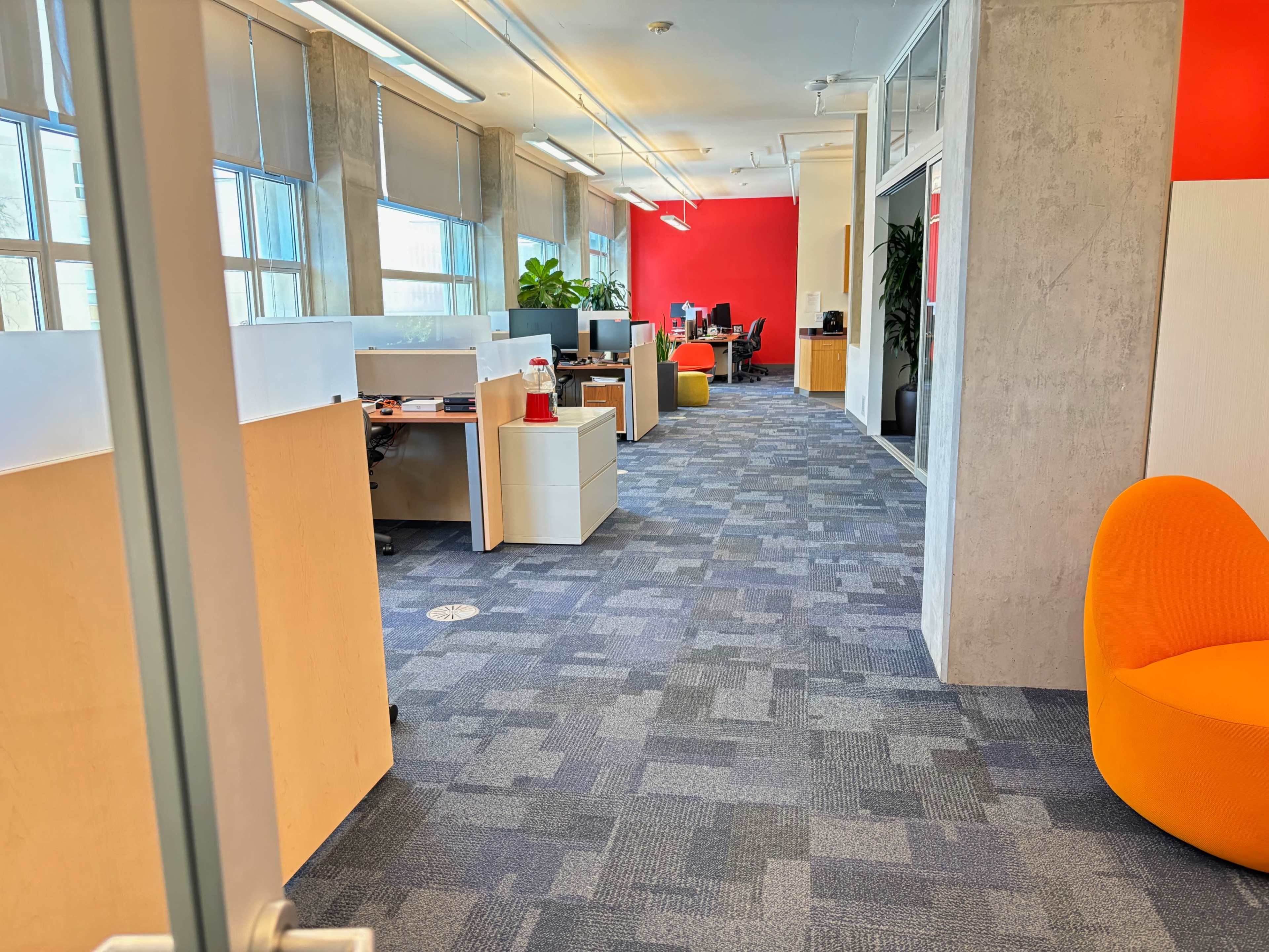 An office space with several workstations, equipped with computers, and orange chairs, along with large windows allowing natural light.