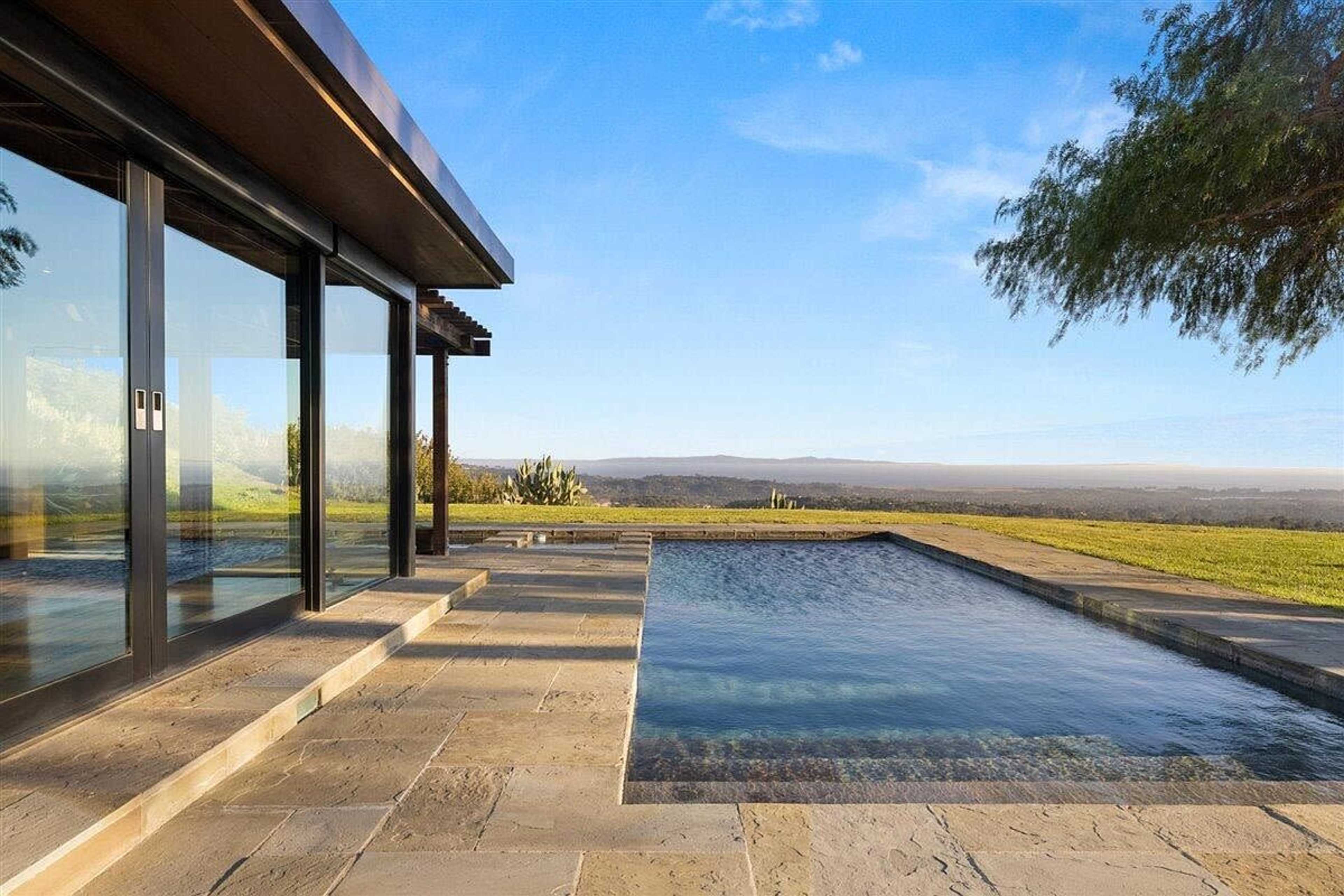 A modern home with large glass doors overlooks a rectangular swimming pool and a valley beyond.