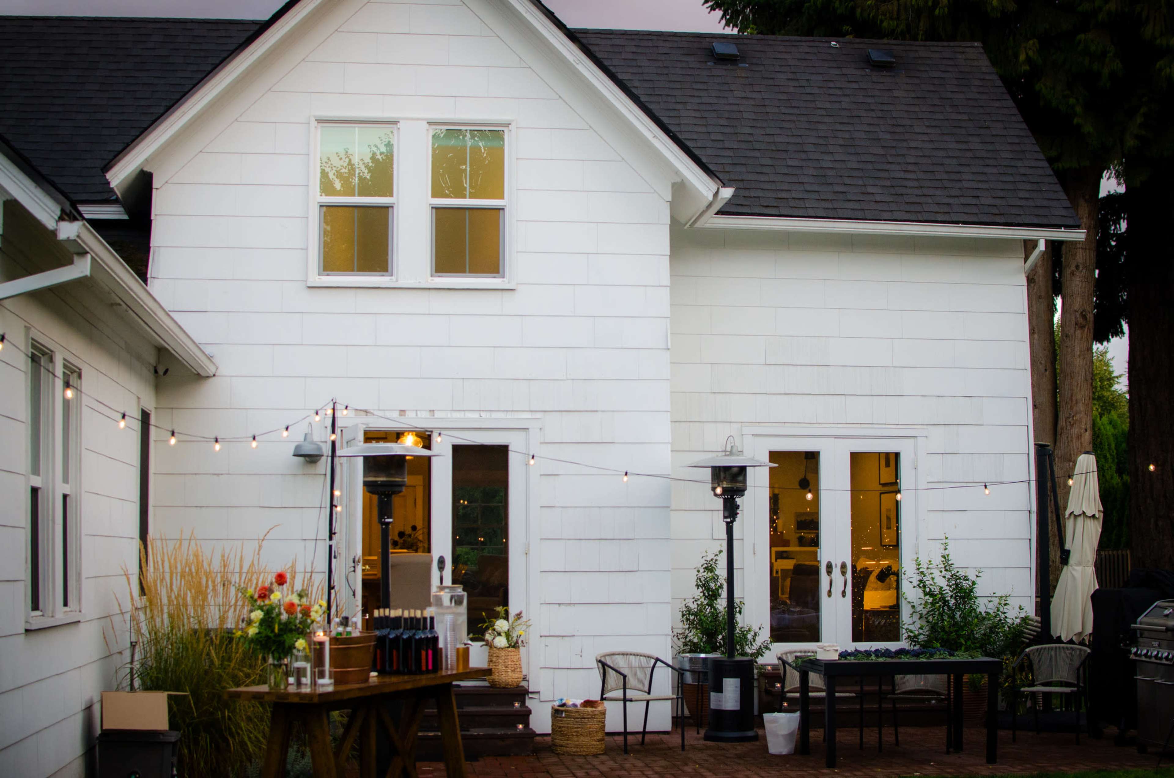 A white two-story house with a backyard setup featuring a wooden table, patio seating, and string lights.
