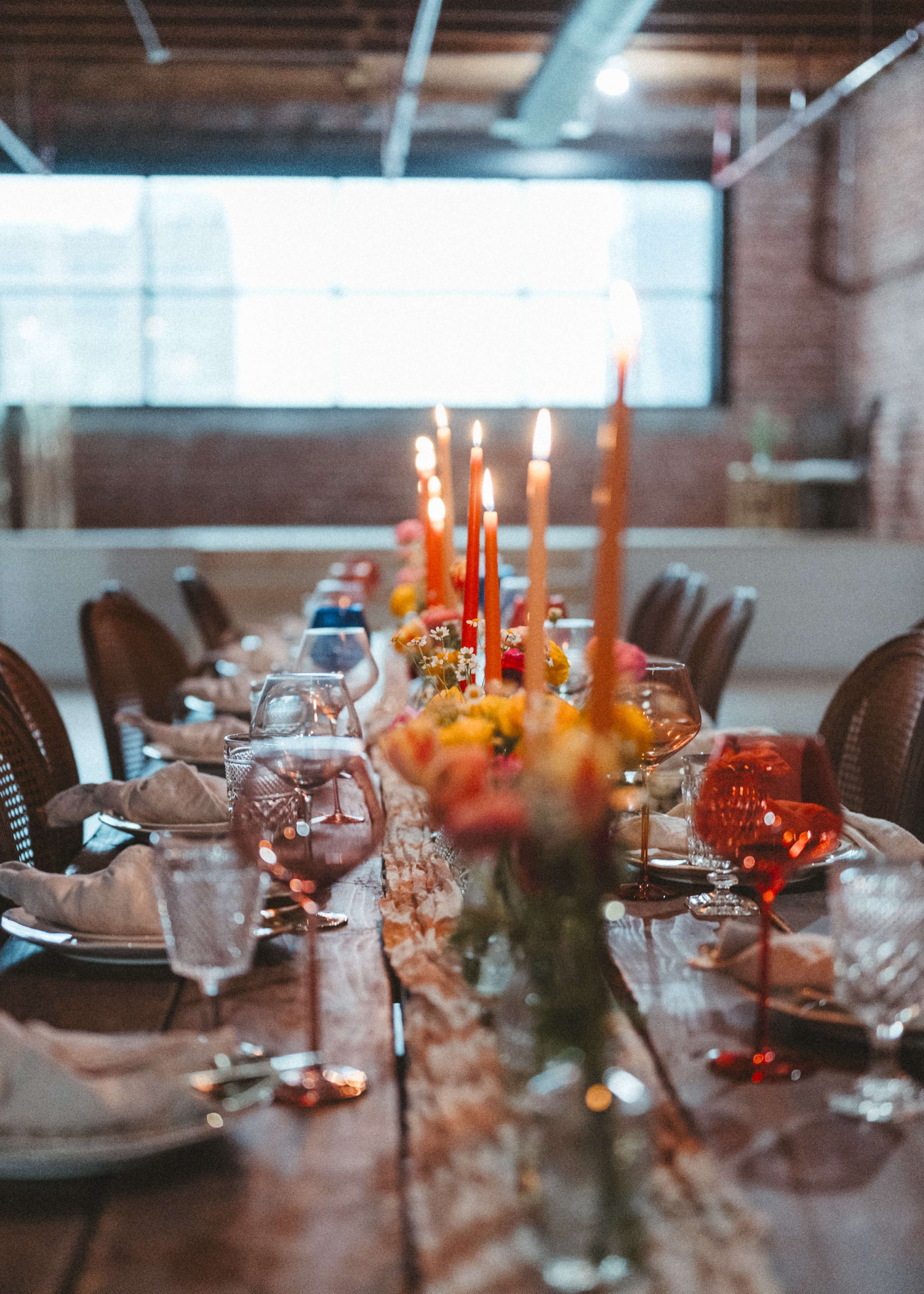 A long wooden table is set with colorful glasses, candles, and a floral centerpiece in a well-lit room with large windows.