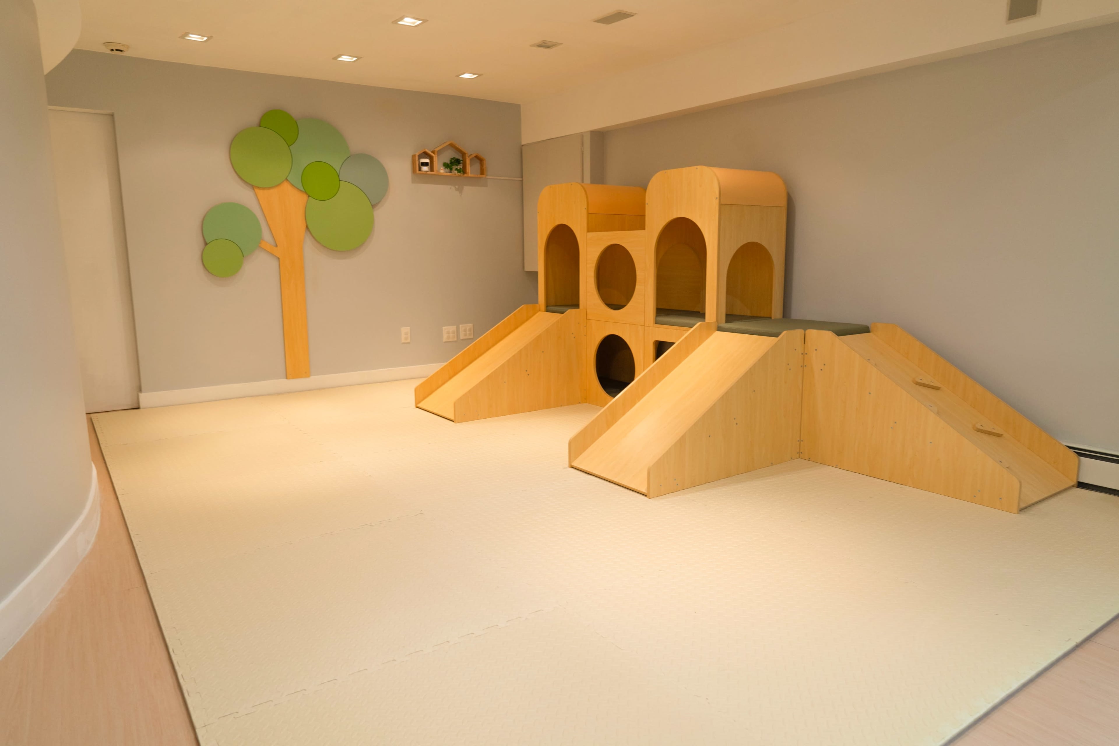 The image shows a playroom with two wooden play structures resembling small hills and a tree mural on the wall.
