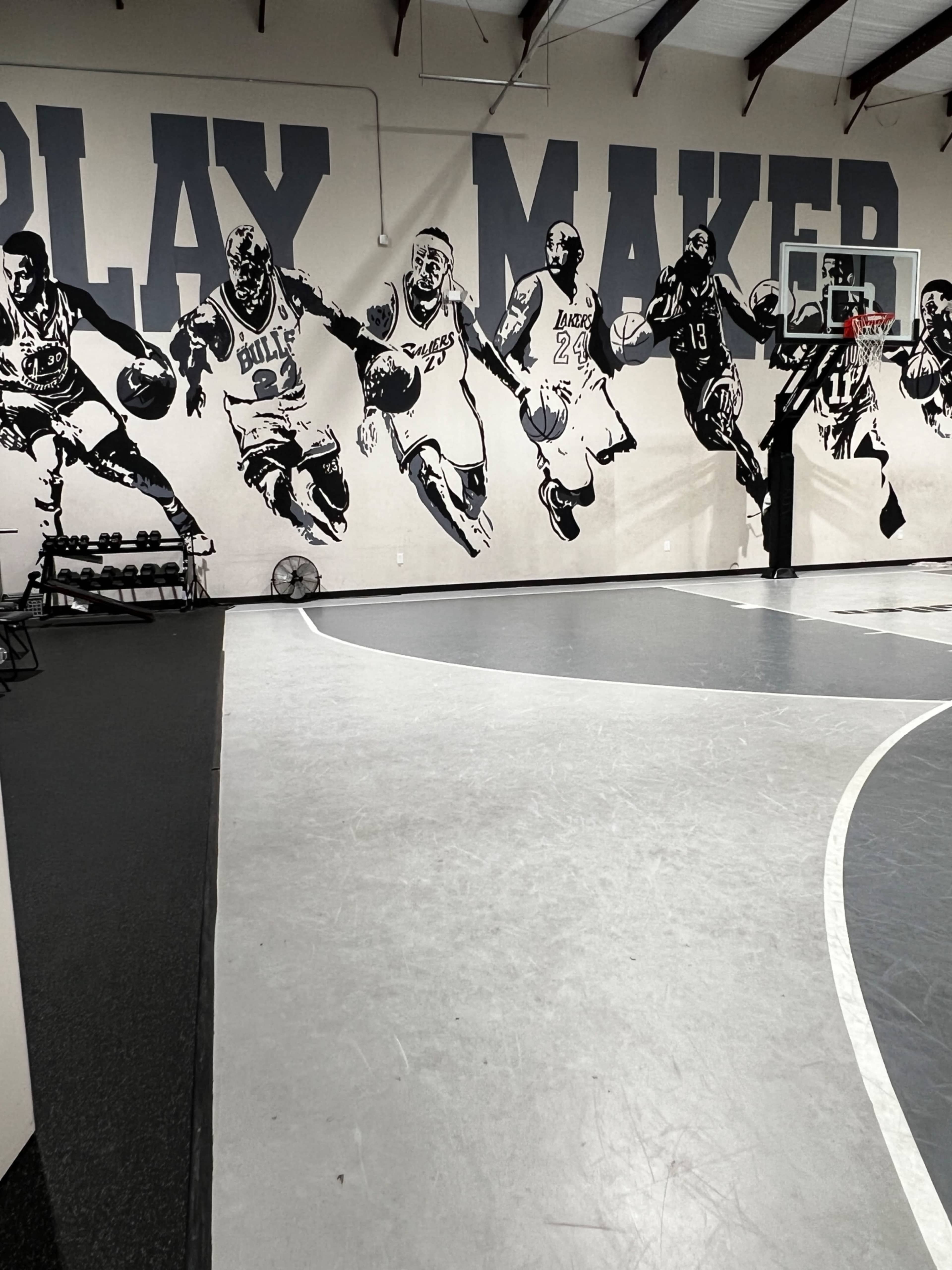 The image shows an indoor basketball court featuring a wall mural of various basketball players in action, with a basketball hoop visible at one end.