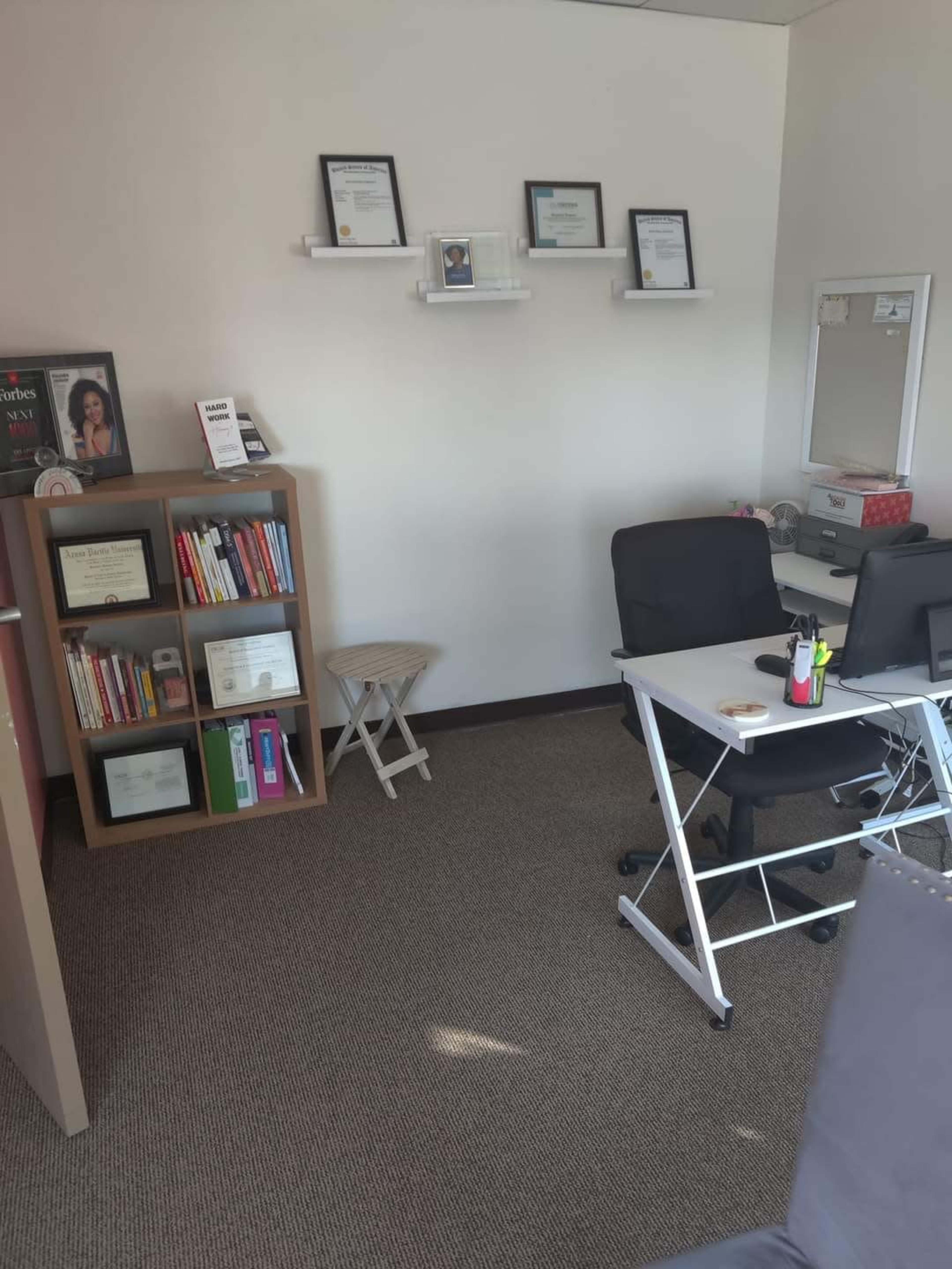 A small office space featuring a bookshelf filled with books and certificates, a white desk with a computer, and a chair near a mirror on the wall.